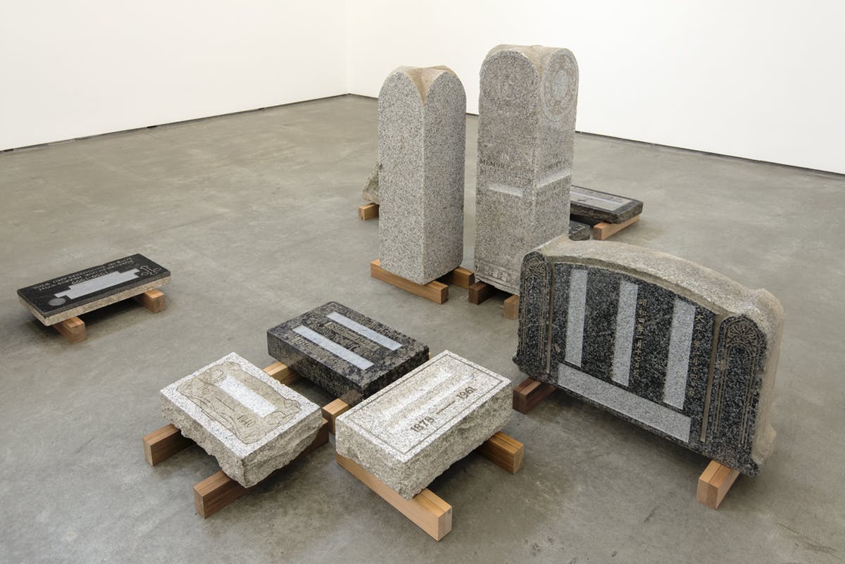 Variously sized tombstones installed in a gallery. A pair of tall, narrow grey stones stand in the middle of the group of tombstones. No names are carved onto these stones.
