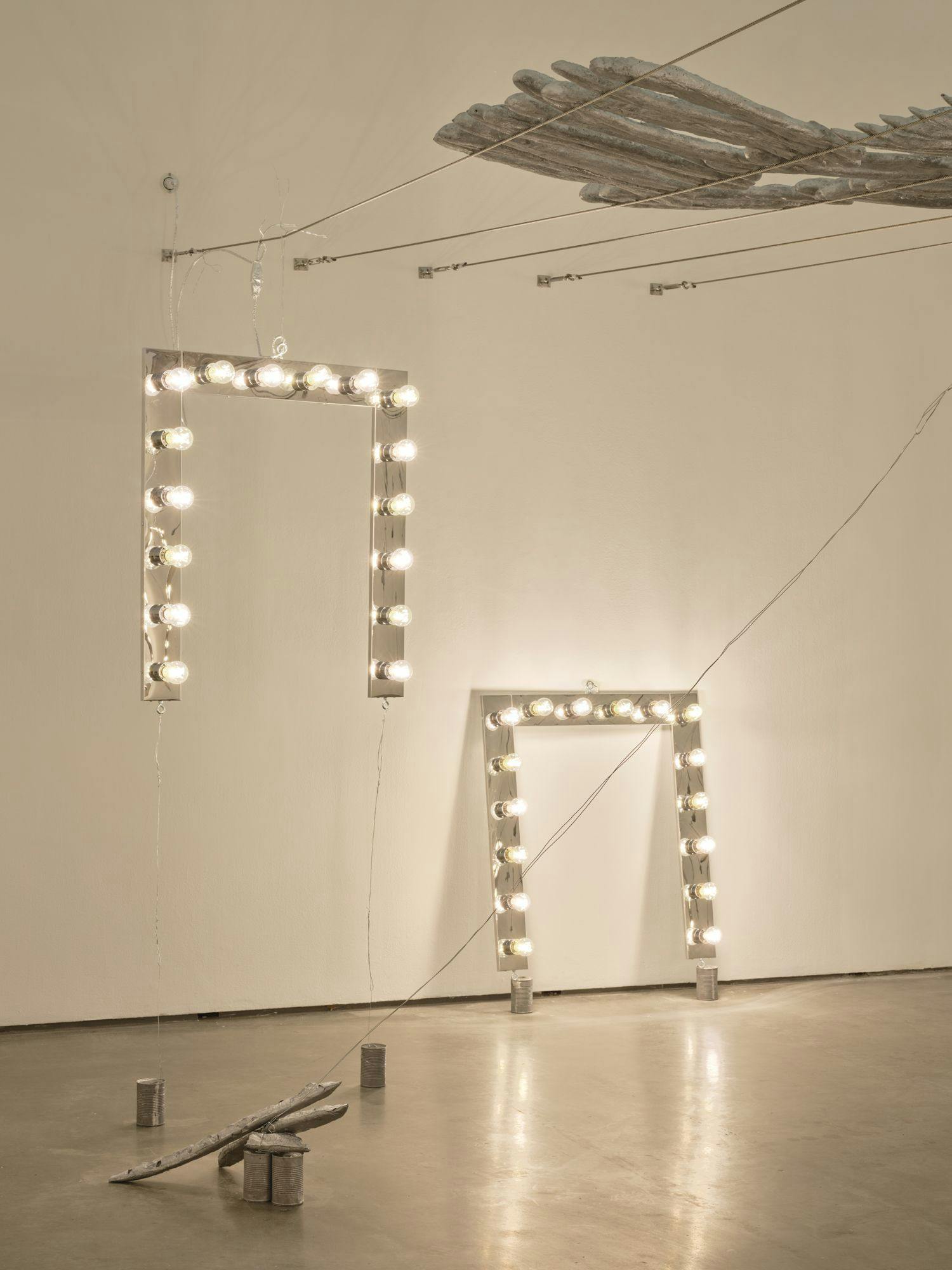 Vanity lights and a dozen silver baguettes hang from wires fixed to the wall. On the floor are another vanity light, more silver baguettes and aluminum cans.