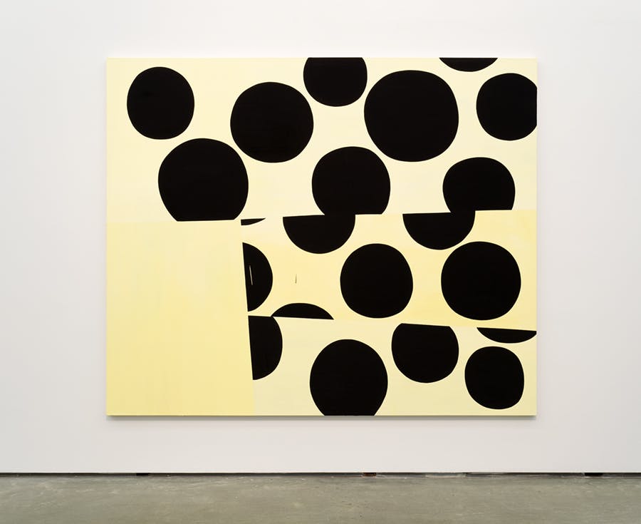 A large-scale artwork mounted on a gallery wall. On top of a light yellow background, groups of large, black, circular shapes are stacked on top of each other in three subtle sections.