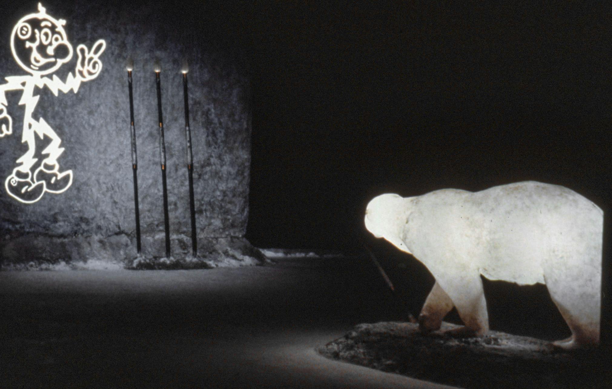 A polar bear sculpture in a dark room is placed to appear to be walking towards a faw stone wall. The wall has a glowing, smiling cartoonish character on it, as well as 3 large spears resting on it. 