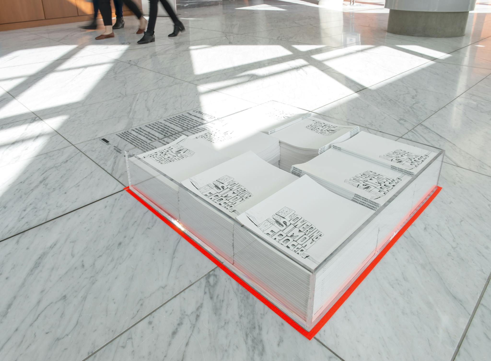 A clear display case with multiple stacks of books sits directly on a marble floor. The book cover reads “Lowering Simon Fraser” in illustrated text. The case is outlined with neon orange tape. 