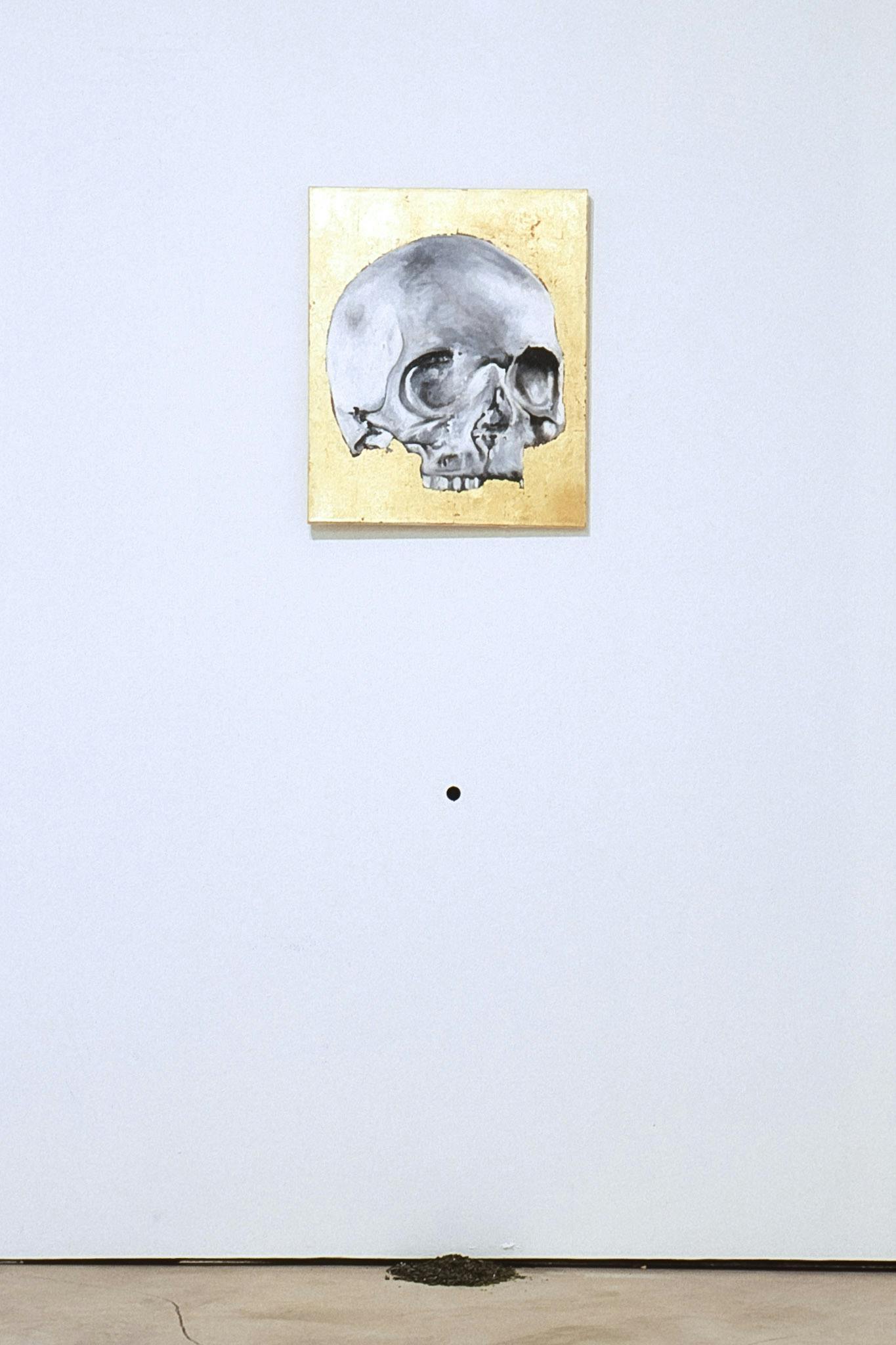 A small gold painting, foregrounded by a silver skull, is mounted on the gallery wall. A small hole is made on the wall beneath where the painting is hung. A dirt pile is on the floor under the hole.