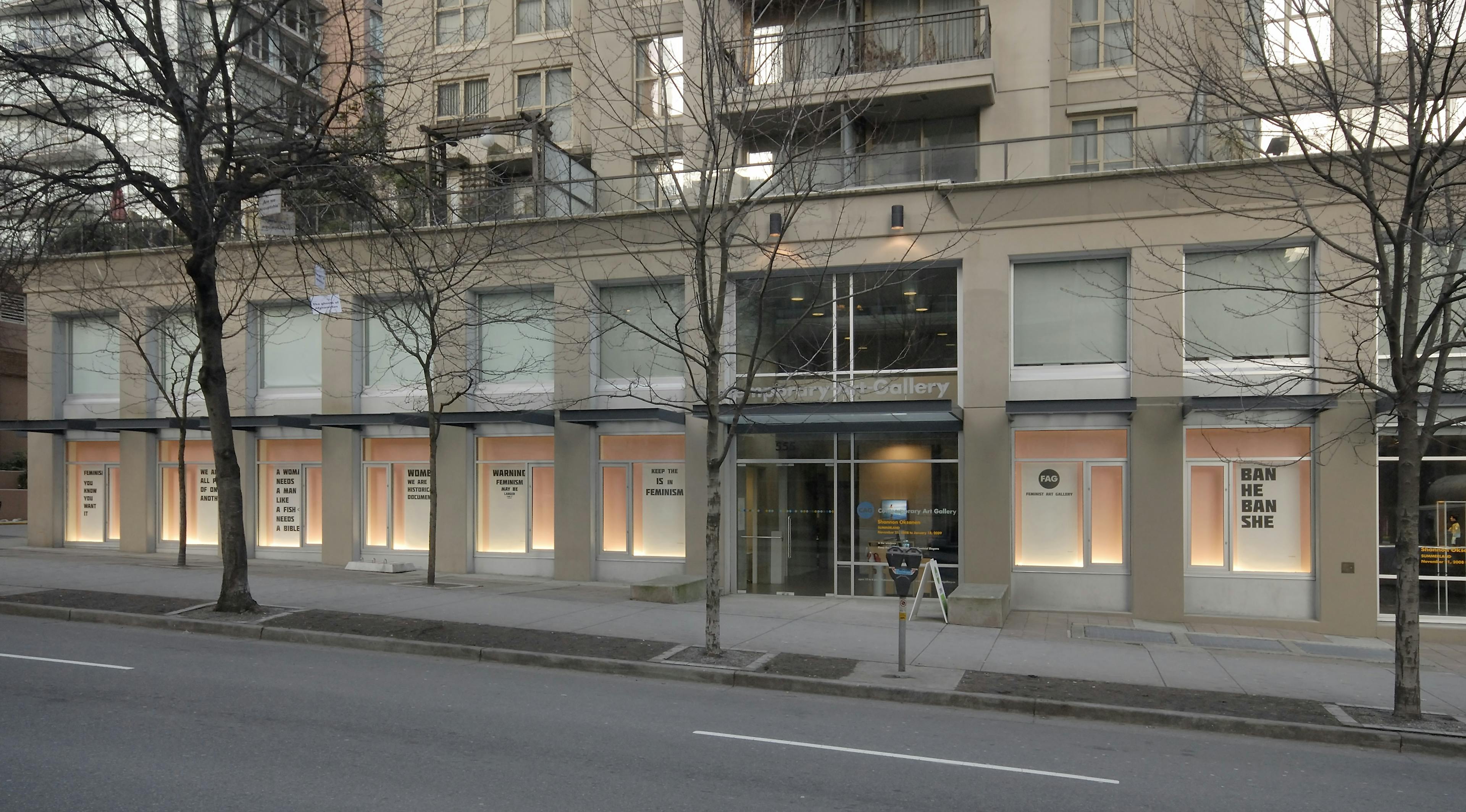 A street view of the CAG window spaces, which are painted pink and each contain a large poster with large black block text. 