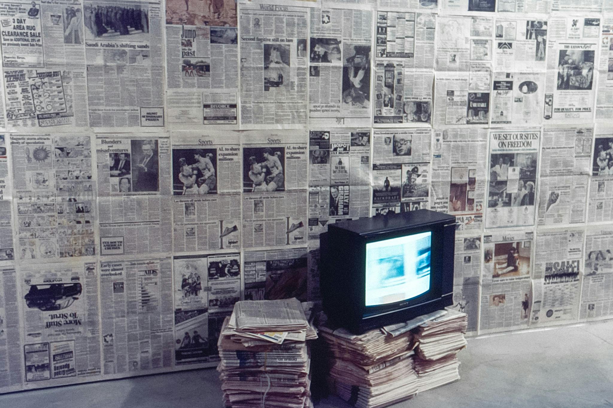 The wall of a gallery space is covered by pages of newspaper. At the base of the wall, there are three stacks of newspapers. A black rectangular tv sits on two of the stacks, playing a blurry video.