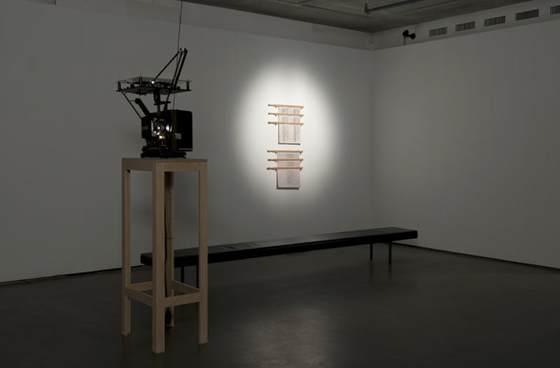 A film projector is installed in the middle of a gallery on a tall wooden support. A text-based artwork is mounted on the wall behind the projector.