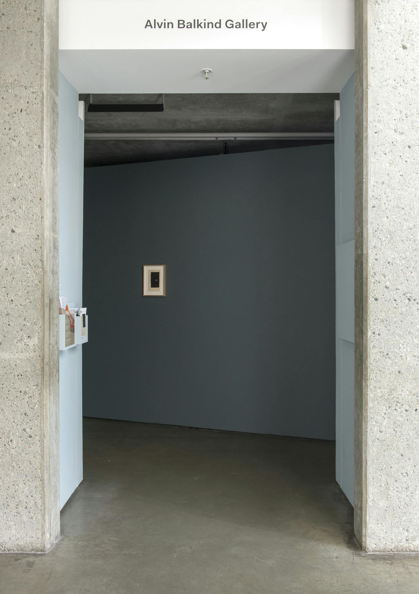 A short hallway leading into a gallery is depicted in this image. A blue-grey wall is visible at the end of this hallway. A small, two-dimensional framed artwork is installed on this wall.
