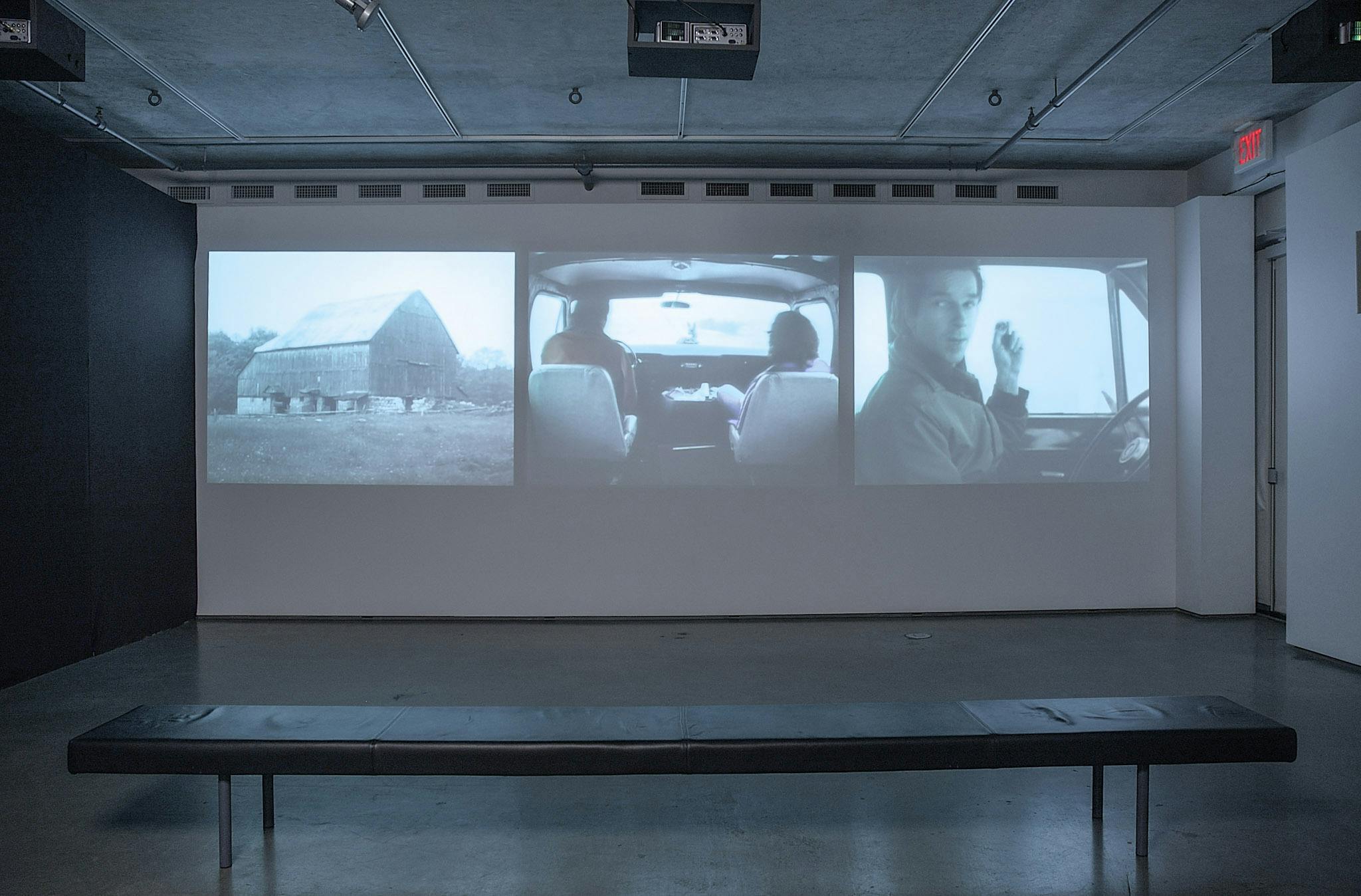 An installation image of three-channel video work projected on a gallery wall. The left video show an image of a longhouse. The middle and right videos show the images of people in a car. 