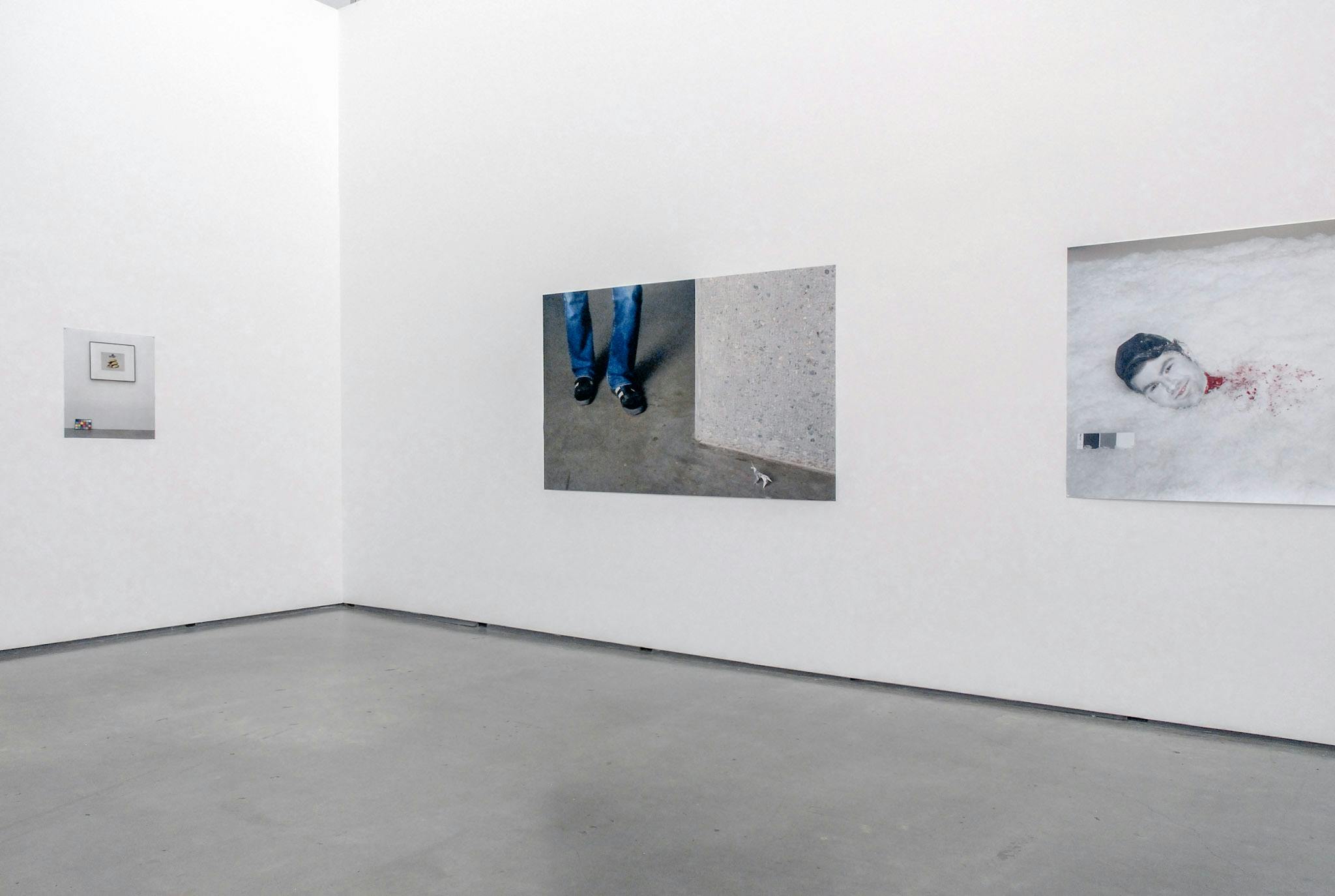 Three photographs on the gallery walls are visible in this image. The photo in the middle shows a person’s feet standing at the corner of a room. The person wears a pair of blue jeans. 