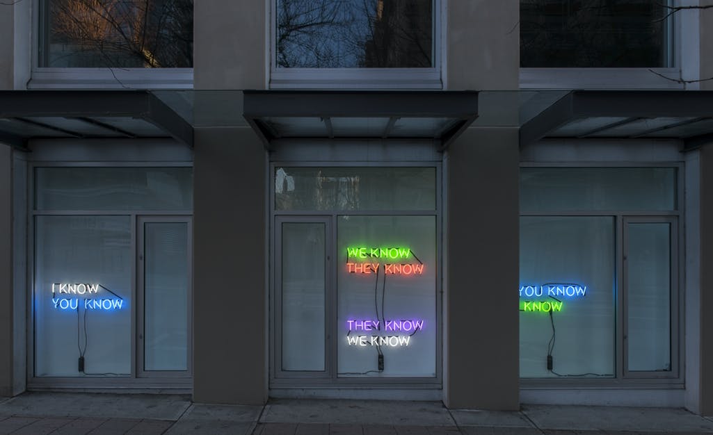 Image of Tim Etchells’ neon text works installed in CAG’s facade windows. Phrases in neon lights of various colours read: “YOU KNOW I KNOW,” and “THEY KNOW WE KNOW.”