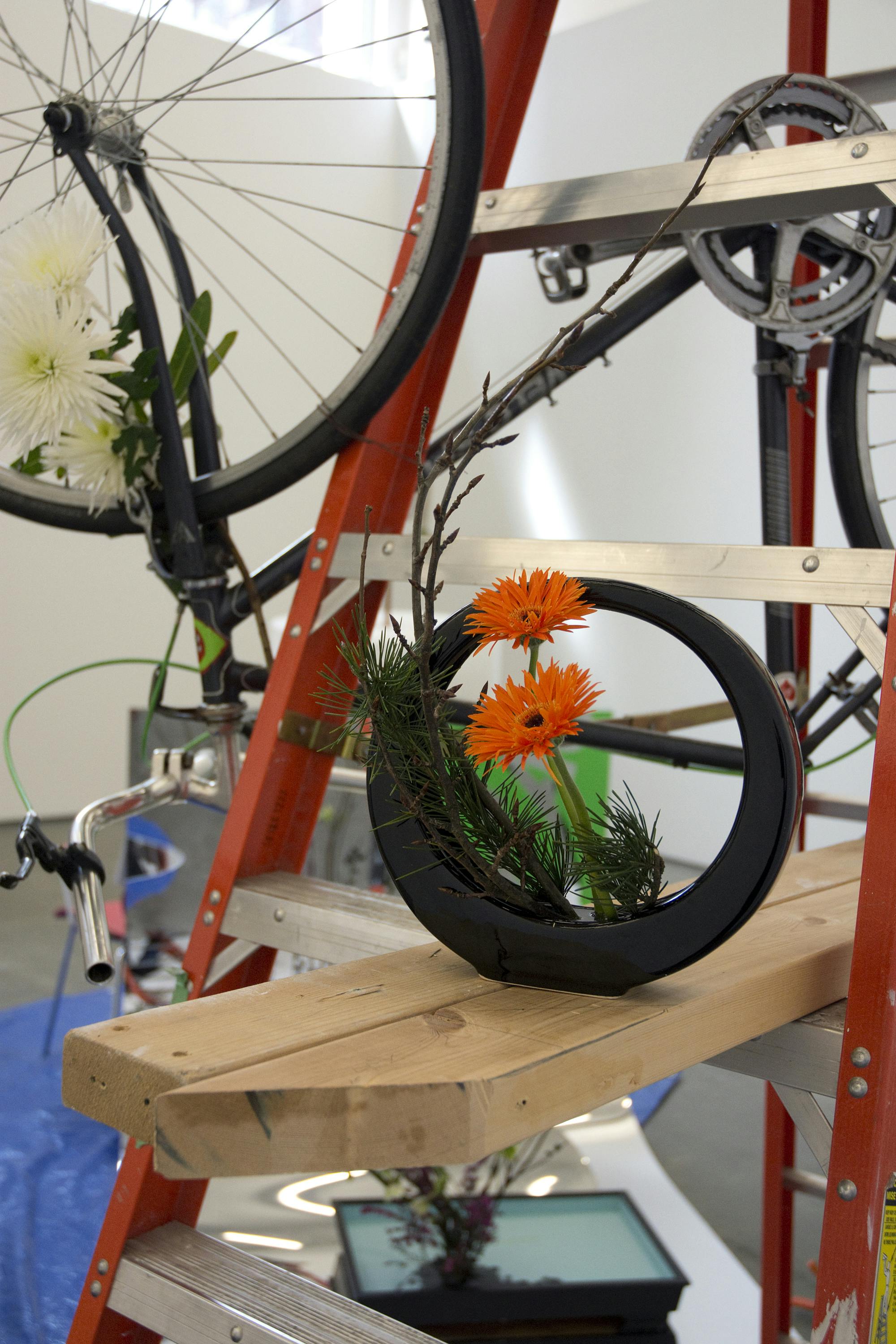 Detail of a sculpture installation. Orange gerberas and small twigs are arranged in a small black ikebana flower vase. The vase is situated on wood bars placed on a step ladder.