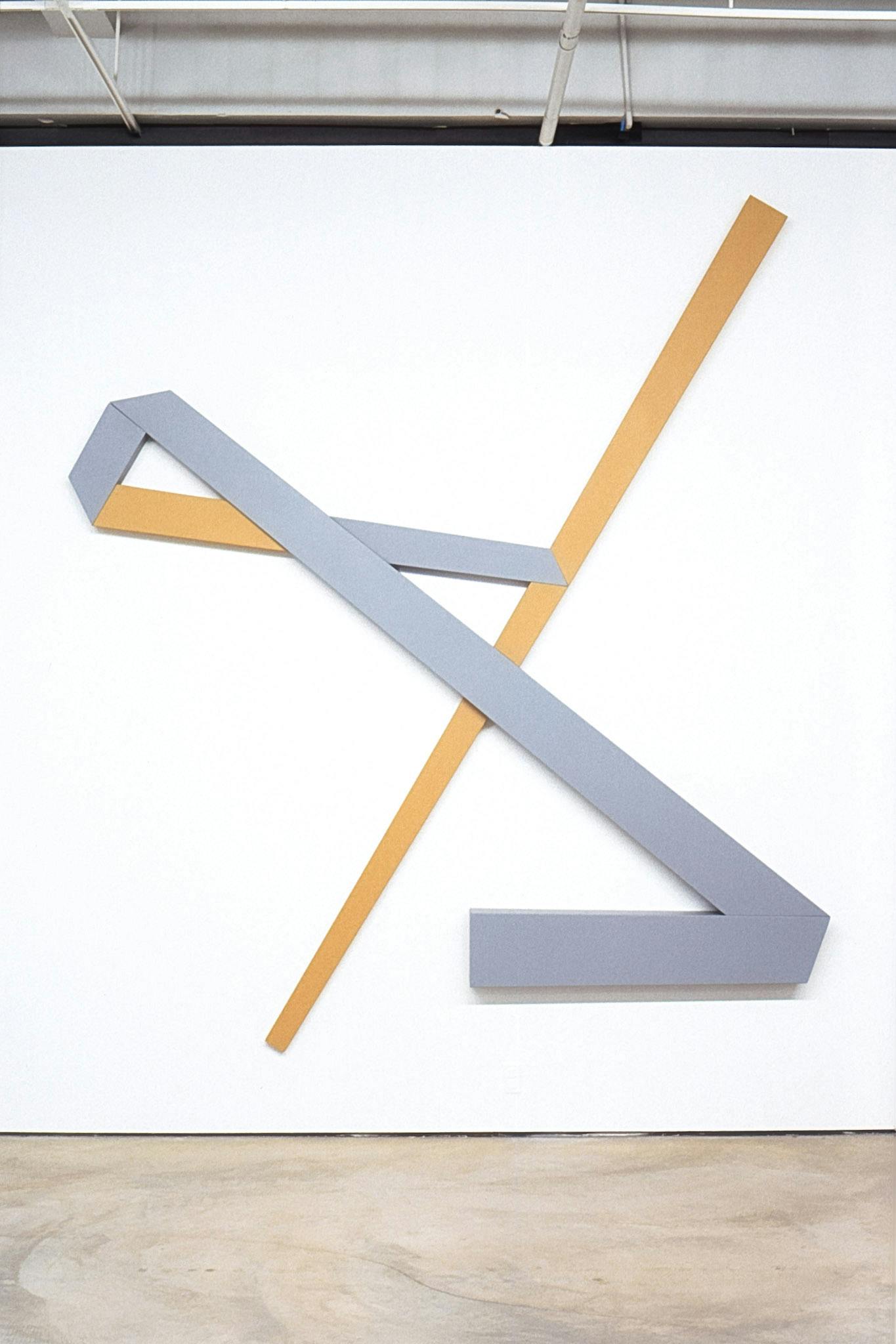 A large-sized geometric sculpture is installed on the gallery wall. A yellow diagonal line runs from the top to the bottom of the wall. This line is foregrounded by an inverted Z shape in light grey.