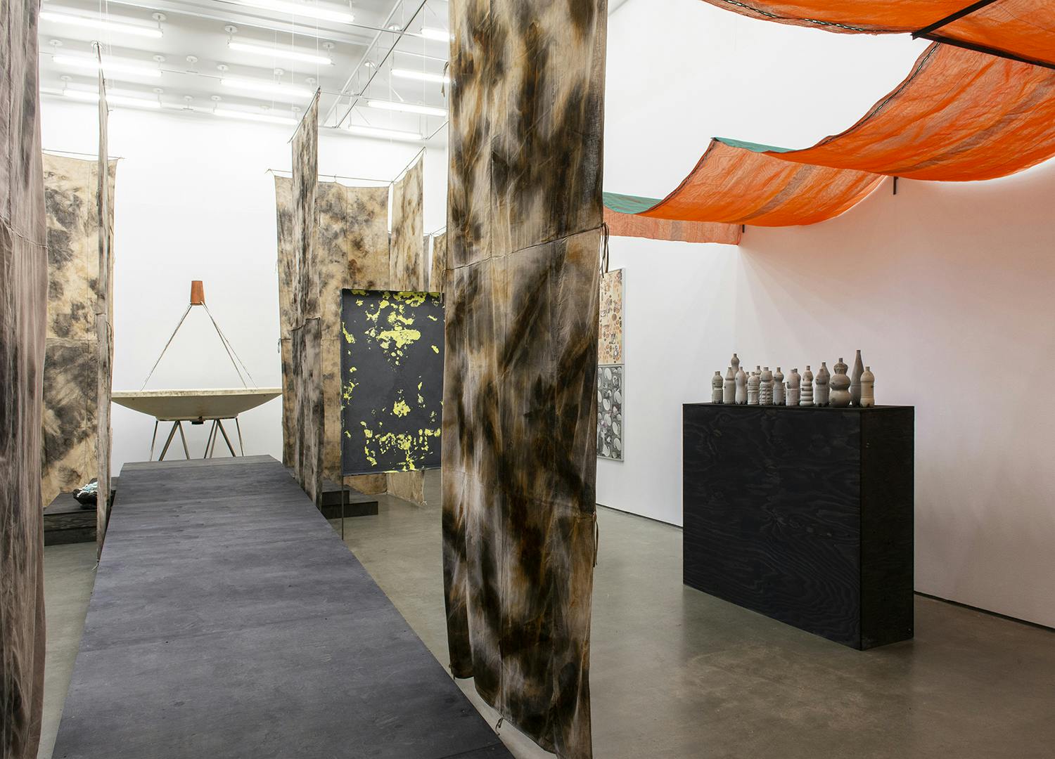 Strips of dyed cloth and orange netting hang throughout the gallery space. To the right a ramp leads to a satellite dish sculpture. On The right side is a black plinth with small sculptures of bottles.