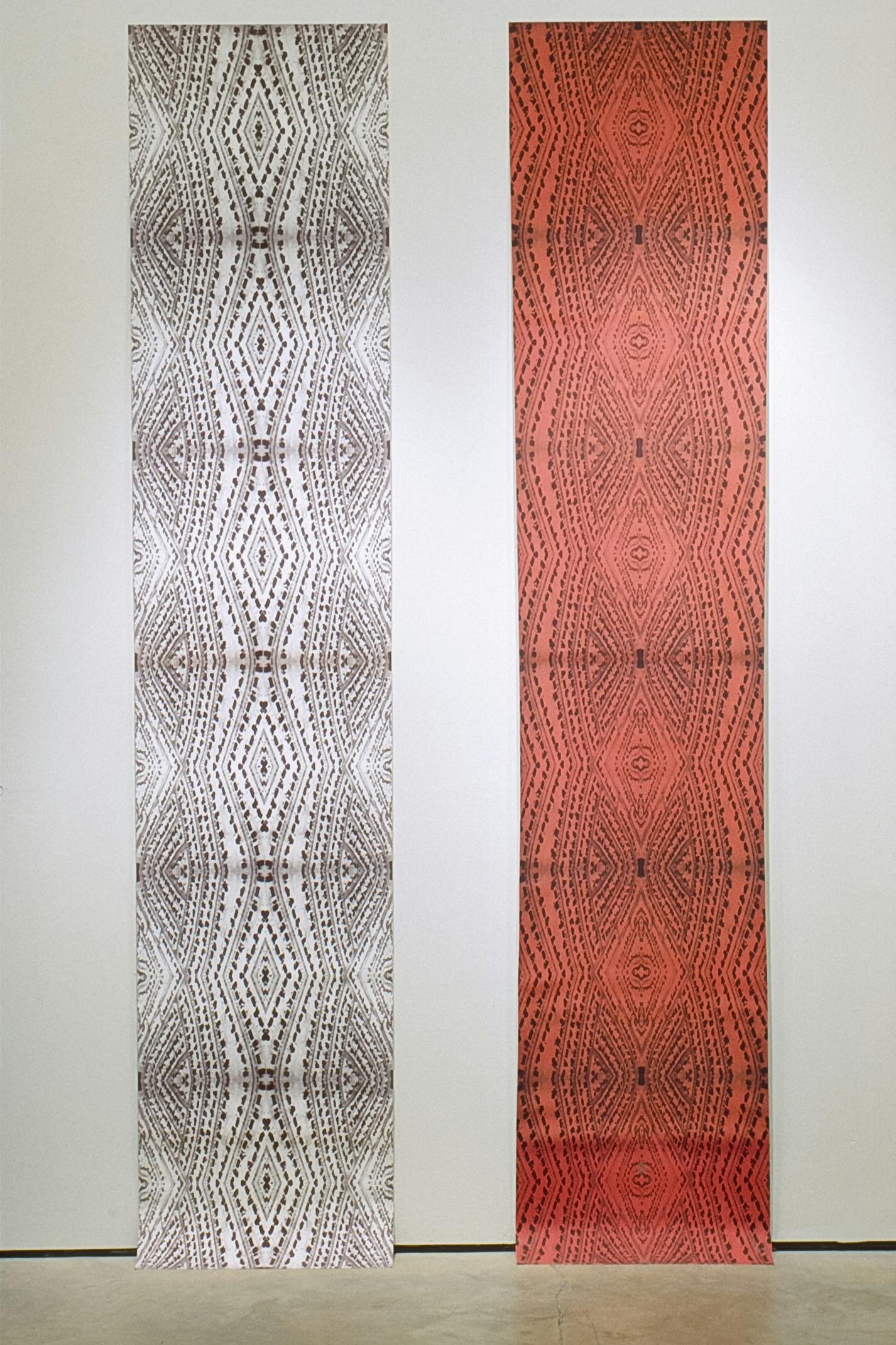 Two scrolls are installed side by side on the white gallery wall. Both have similar intricate patterns printed on the surfaces. The one on the left is white and the one on the right is red.  