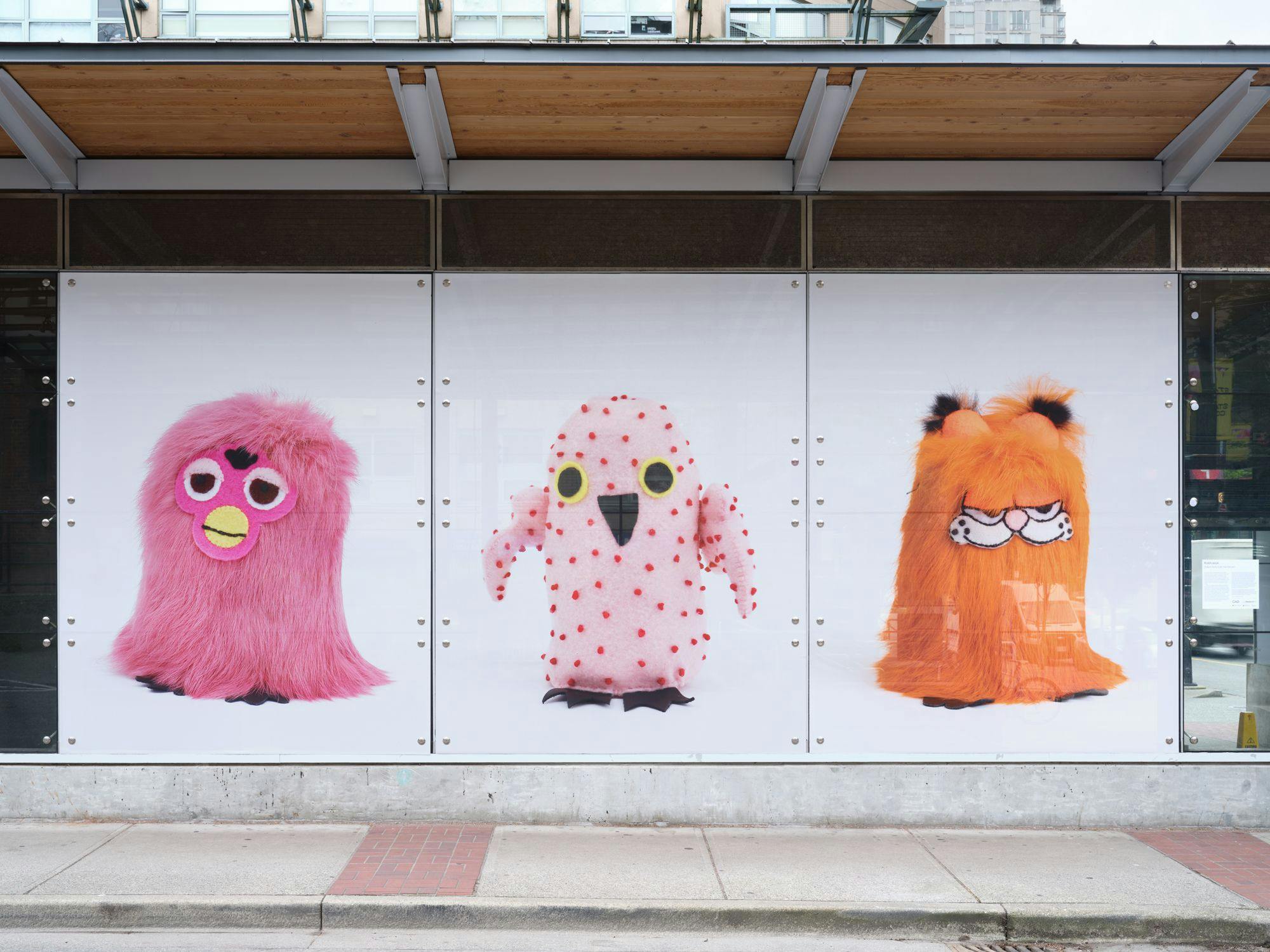 A frontal view of a SkyTrain station with photographs depicting Ookpik figures, or stuffed owl toys, in its windows. From left to right are a Furby-inspired Ookpik, one with no fur and a Garfield-inspired one.