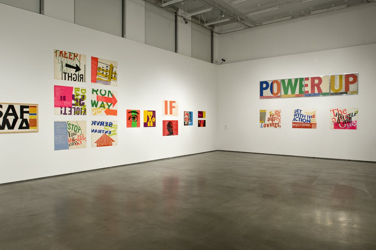 Installation view of Corita Kent’s colourful sign artworks mounted on white gallery walls. Some of those signs read “POWER UP” and “STOP Take it easy. Let yourself Go.”