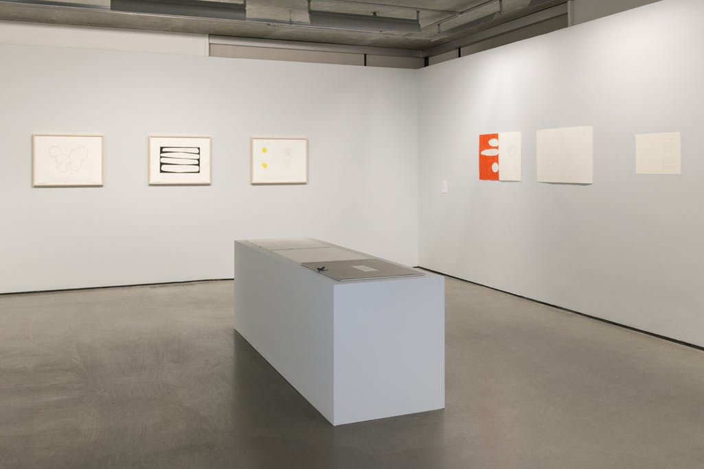 Multiple two-dimensional artworks are installed in a gallery space, including drawings and watercolour paintings. Several pieces are placed on a large plinth situated in the middle of the gallery.