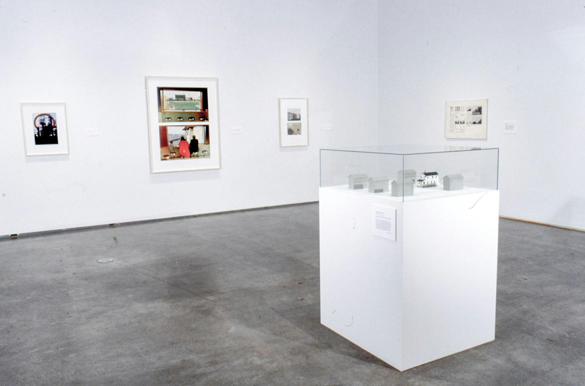 On a large plinth placed in the middle of the gallery, six square-shaped sculptures sit. Four framed two-dimensional works are mounted on the walls around this plinth. 