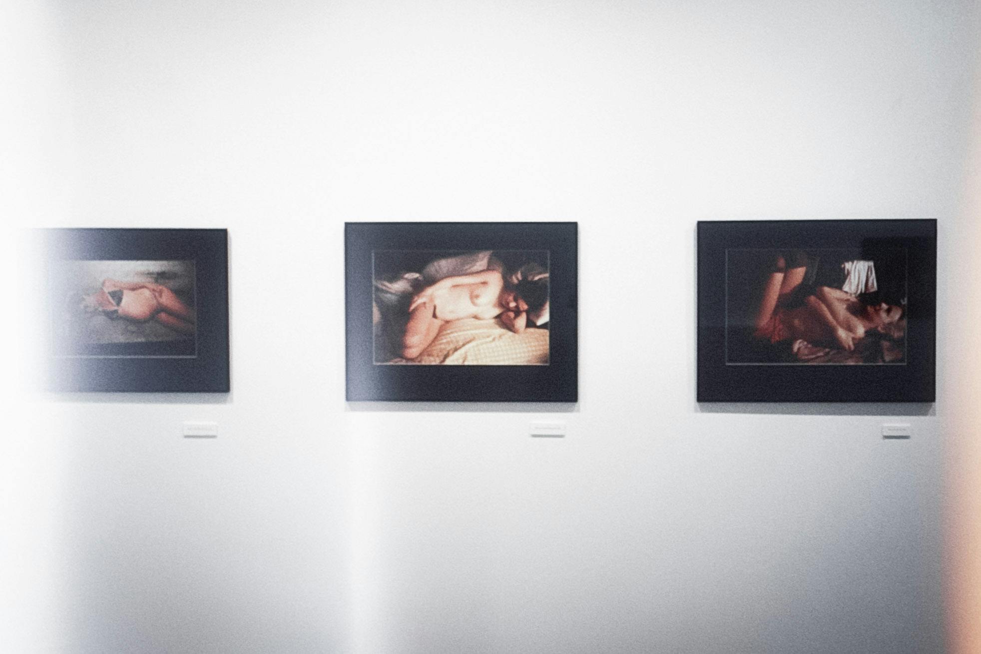 A closeup of three photos in black frames on a white wall. The photos all show people laying down in different stages of nudity. One person is pregnant, one is in bed, and one is sunbathing in shorts.