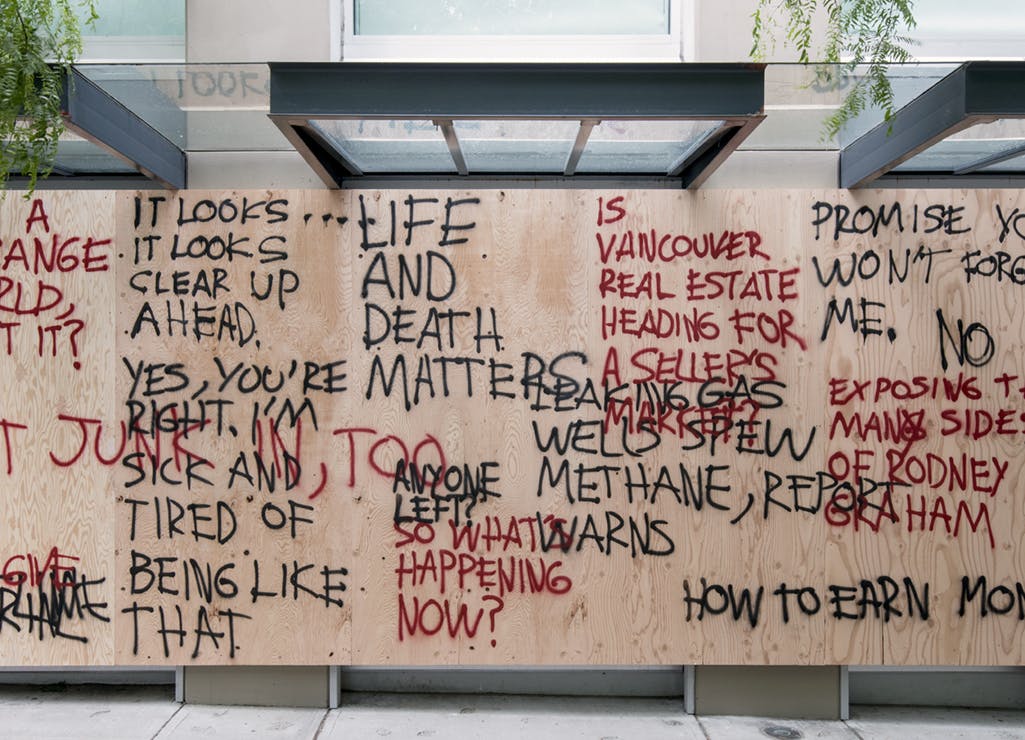 Detail image of plywood boards installed across CAG’s facade windows. Overlapping spray-painted words in red and black cover the boards. One phrase reads, “PROMISE YOU WON’T FORGET ME.” 
