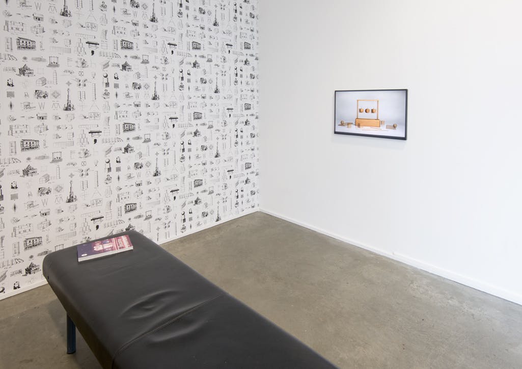 A black bench is installed in a gallery. One wall is covered by a wallpaper with repeating patterns of the artist’s drawings. A monitor displays an image of a wooden object on the other wall. 