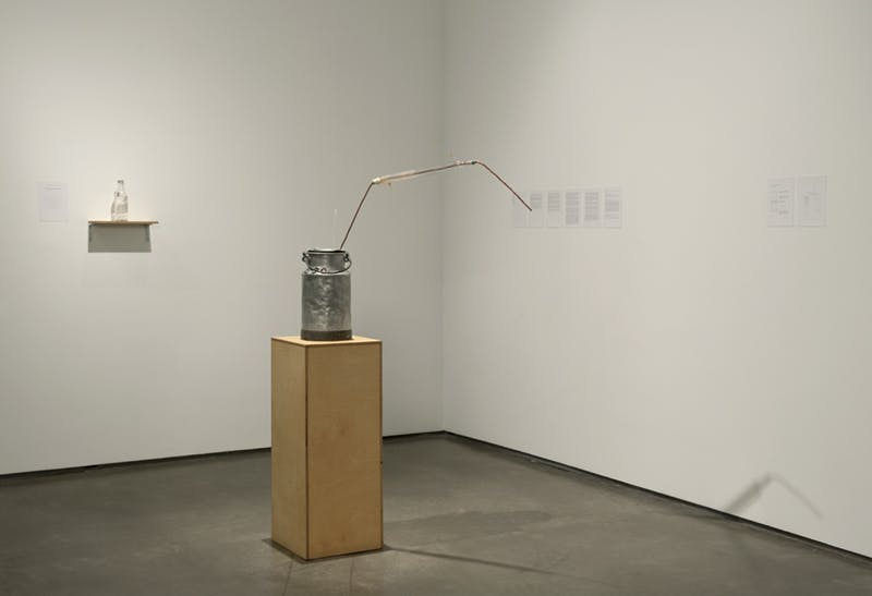 A sculpture resembling a metal milk can sits atop a wooden plinth. A long metal rod, bent at one end, emerges from the top of the jug. A glass bottle is placed on a shelf mounted to the wall.