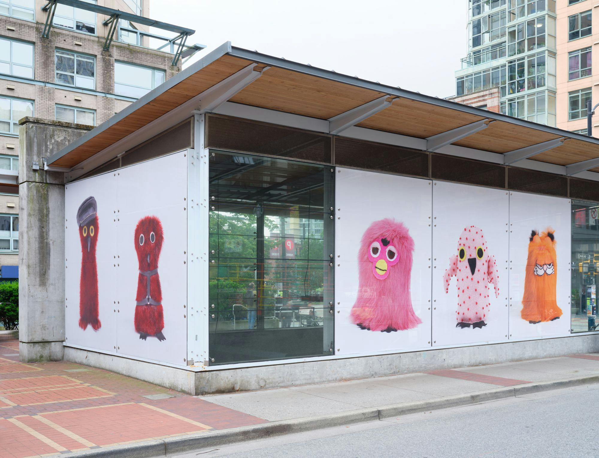 A corner view of a SkyTrain station with photographs of Ookpiit in its windows. From left to right: two Ookpiit with leather gear, a Furby-inspired one, one with no fur and a Garfield-inspired one.