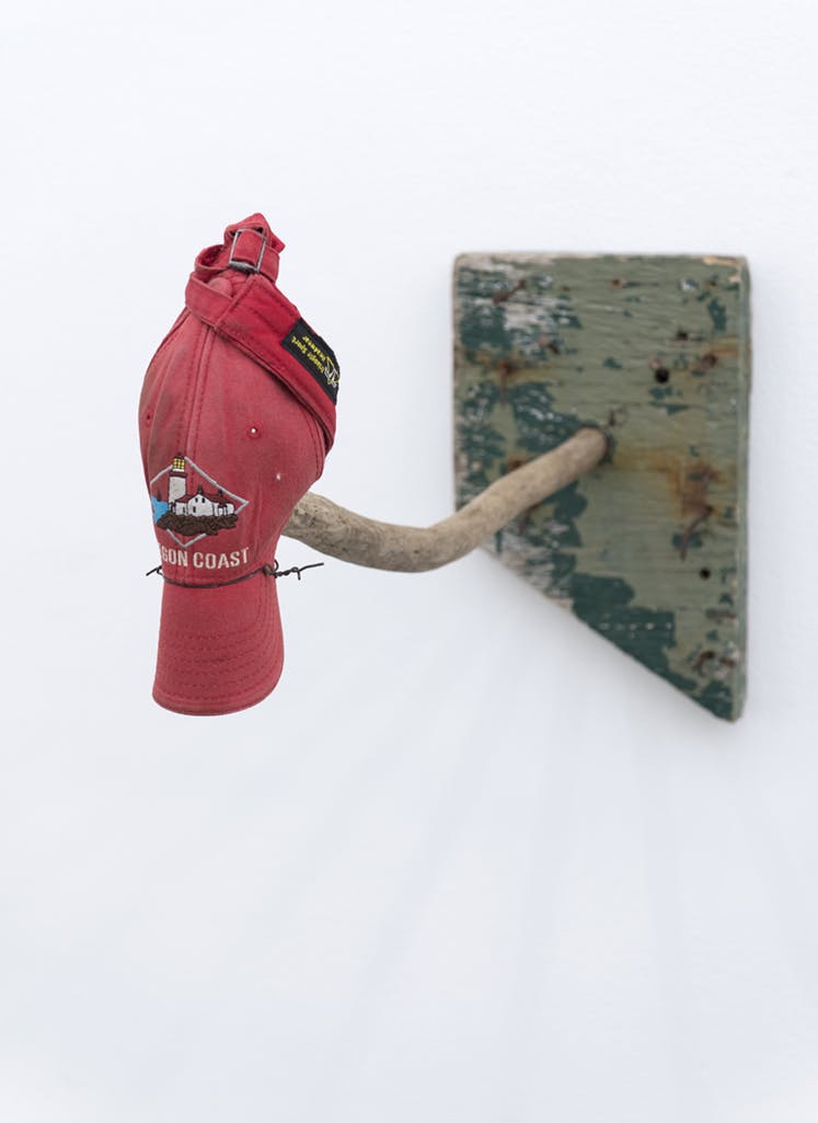 A red cap hangs from a short wood stick installed vertically on a gallery wall. The cap is tied at the back, making its form resemble a bird resting on a tree. 