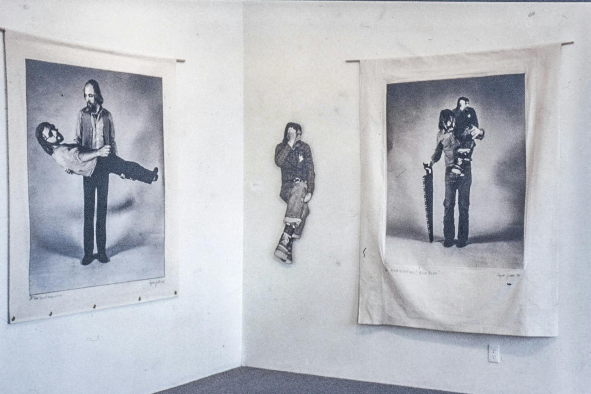 In the corner of a gallery, there are two large photos printed on favribm and a small cardboard cutout mounted on the walls. The photos each show a person with a cardboard cutout of themselves. 