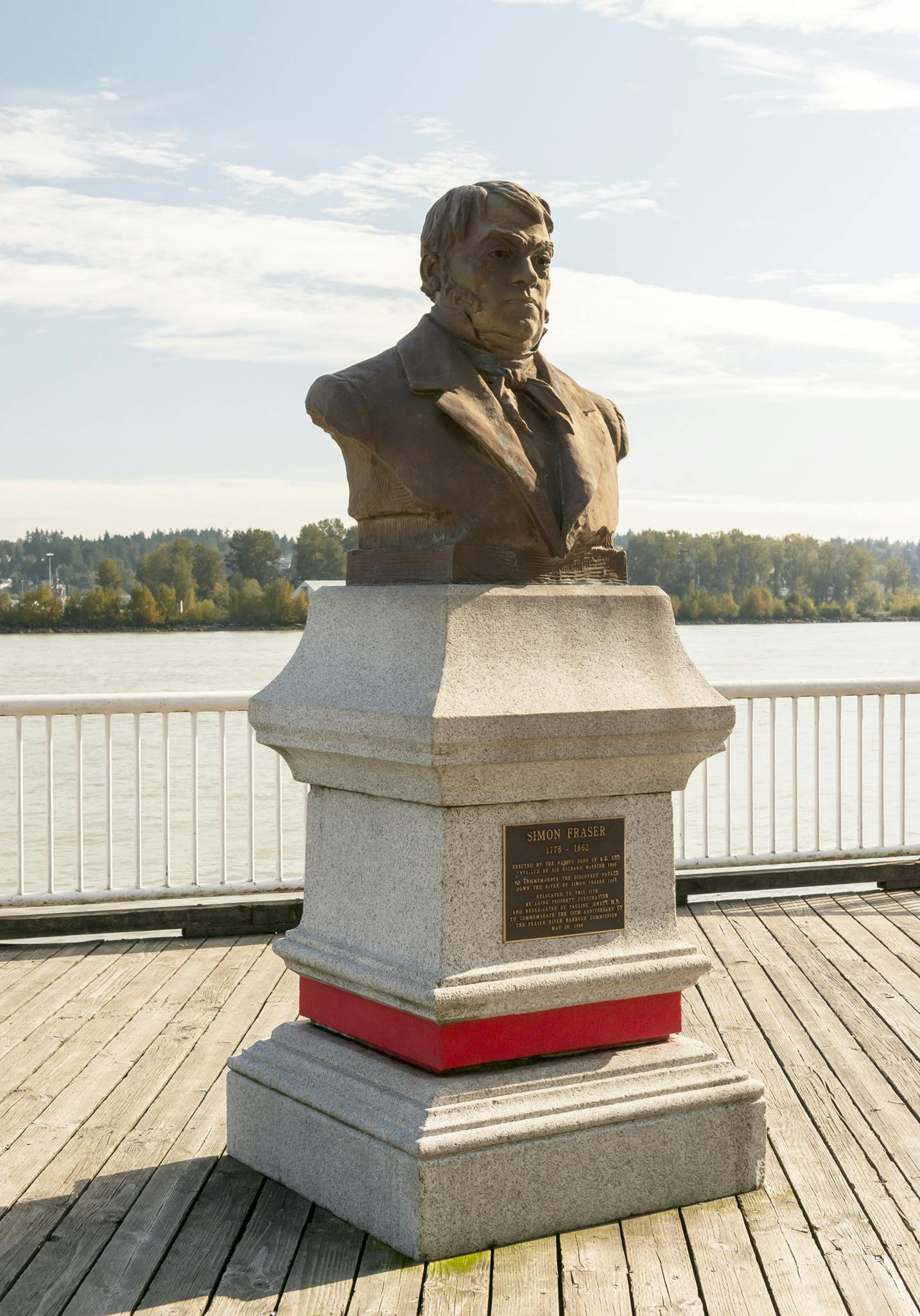 An image of a monument to Simon Fraser installed on a wooden pier by water. The monument is a bronze bust of Fraser atop a granite plinth.
