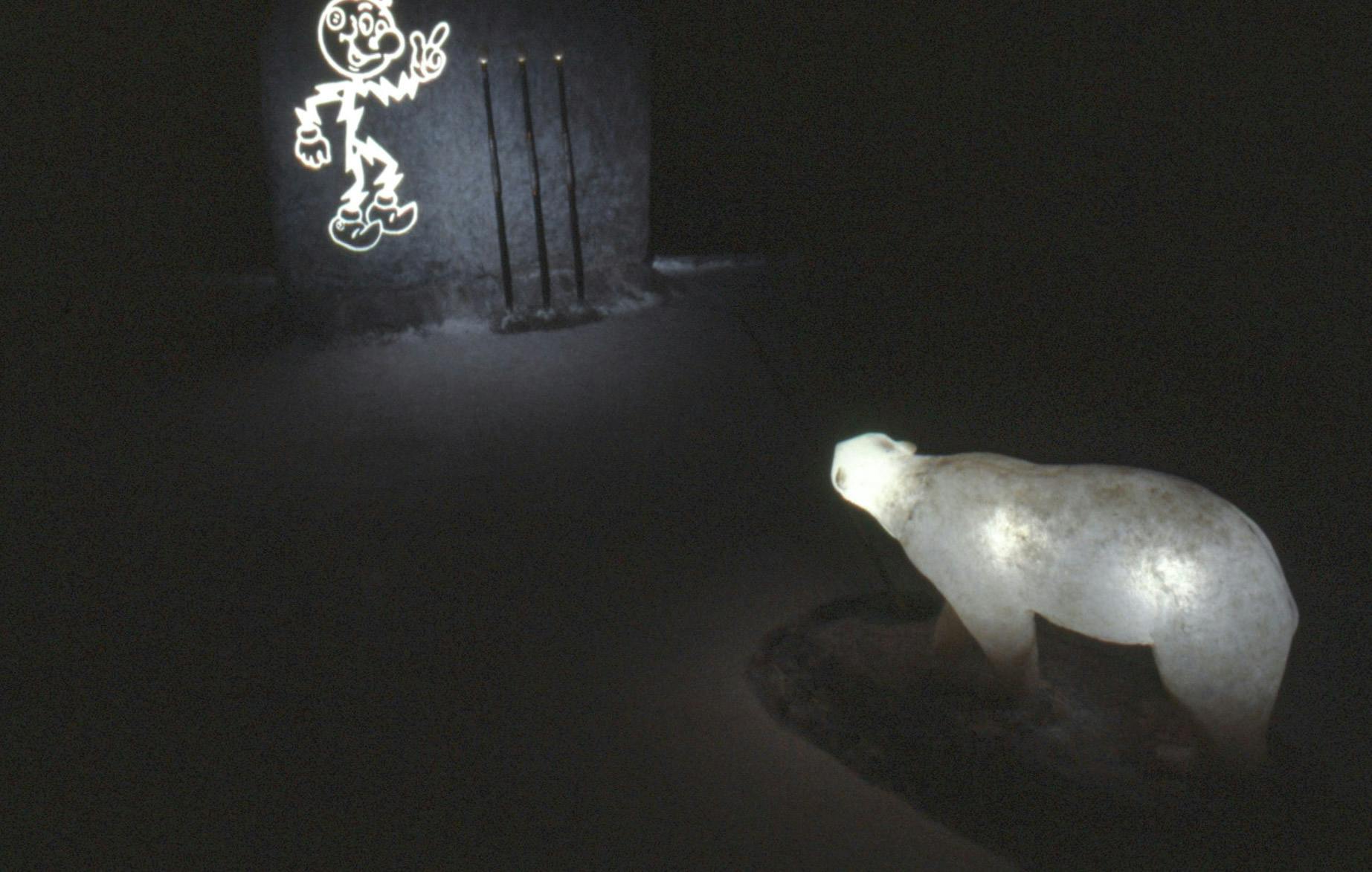 On one side of a dark room, there is a glowing polar bear sculpture, and a faux stone wall with 3 spears resting against it. The wall shows a lit up character with a lightning bolt body on the other.