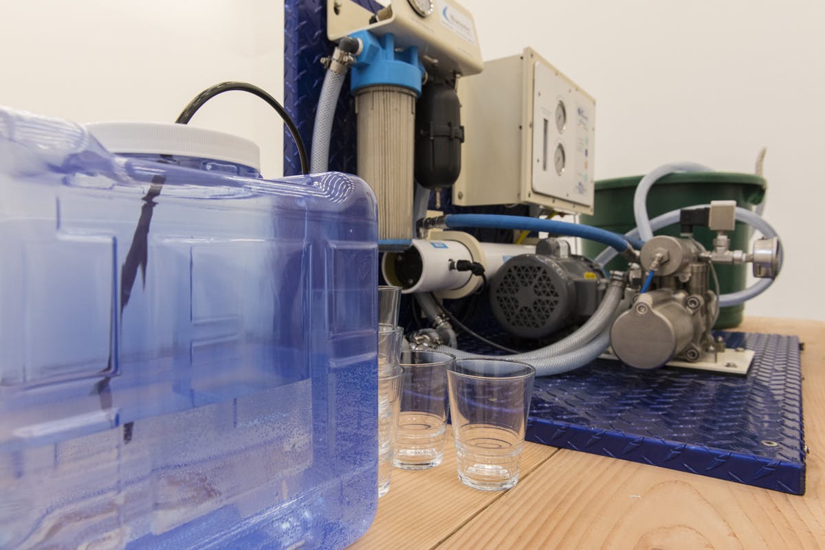 A detail image of objects on a wooden table. A water tank appears next to an elaborate machine sitting on a tabletop. The machine is made of many components of both plastic and metal. 