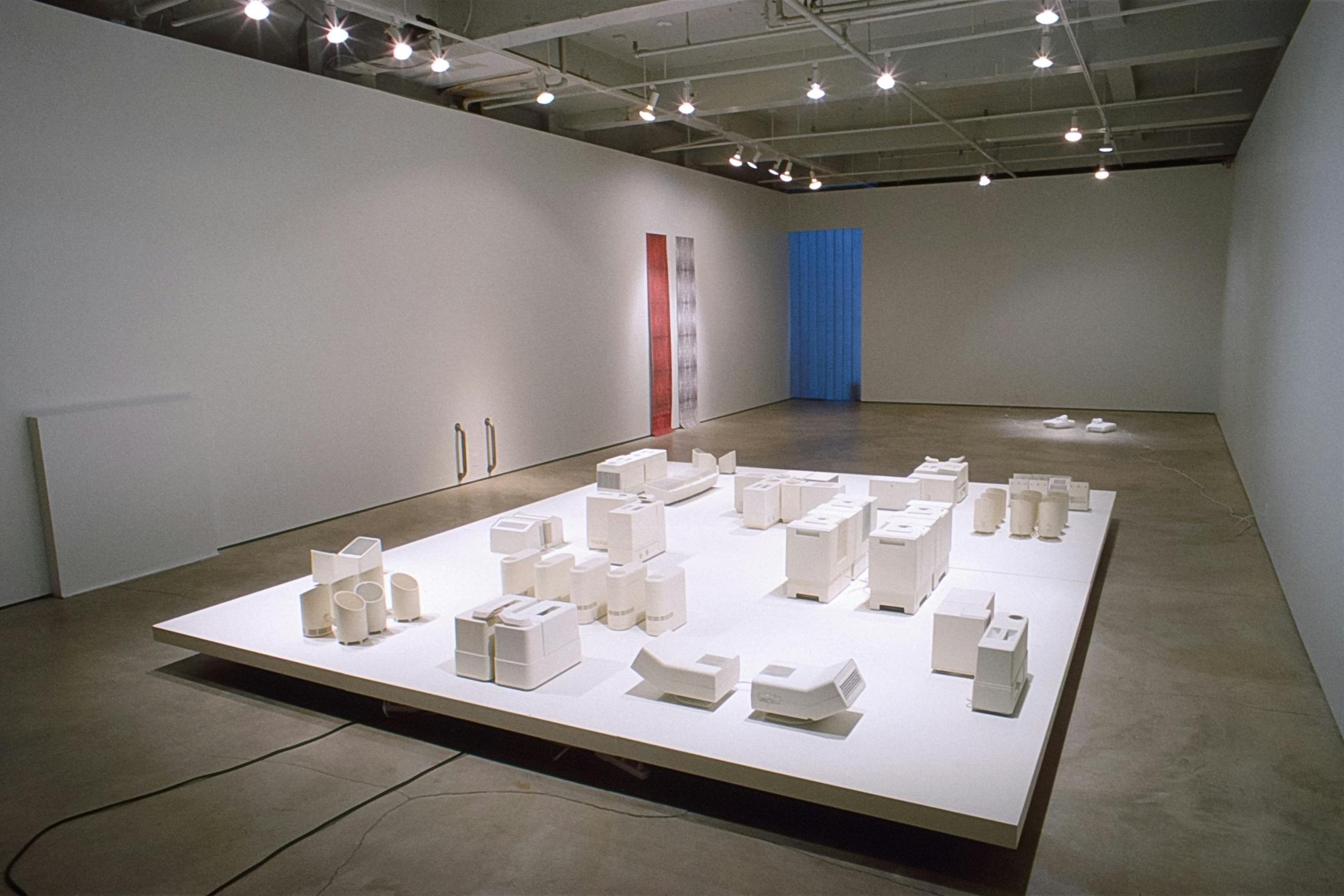 Several artworks are installed in the gallery. White miniatures of electronics are installed on the floor. A pair of red and white scrolls are installed on the back wall. 