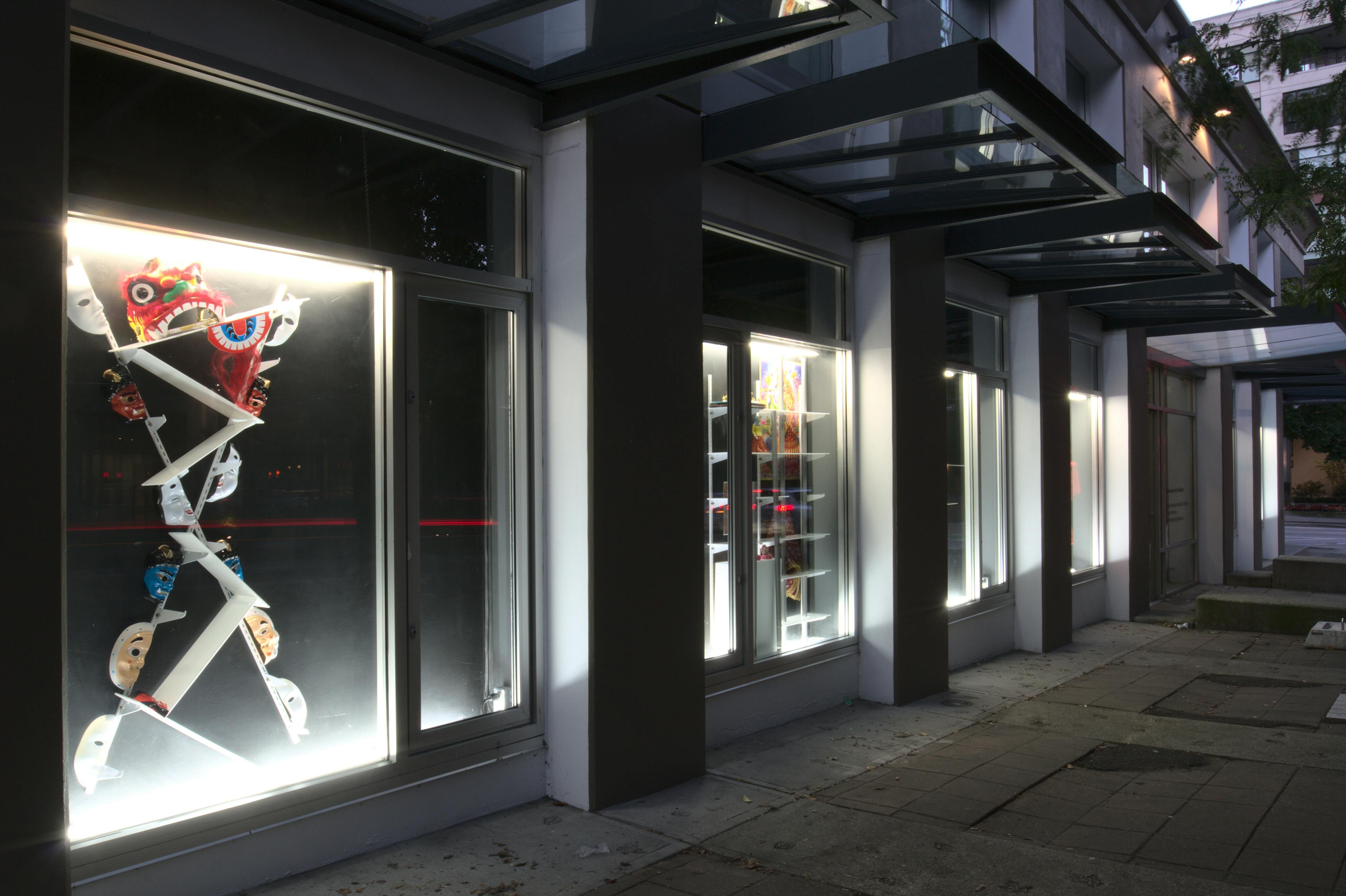 Image of CAG’s exterior facade windows. Installed in one of the windows are masks on a black background. Each mask is split in half and connected to a white, X-shaped shelf and bracket structure. 