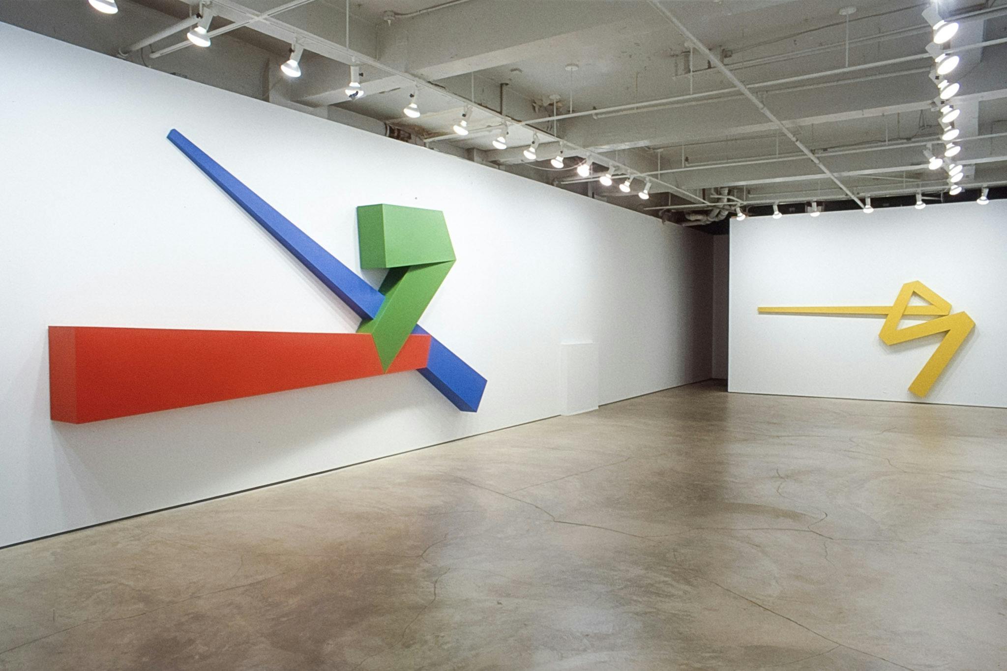 Two geometric sculptures are installed on the gallery walls. The left piece includes figures and straight lines in vivid red, green and blue. The piece on the right is yellow and resembles an arrow.   