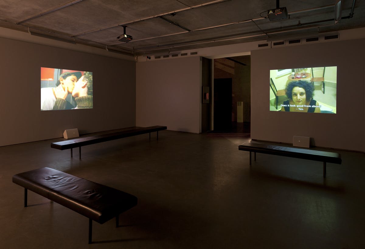 In a gallery space, two video works are projected on the walls. The video on the left shows two people kissing with their eyes closed, and another video shows a person looking up at the camera. 