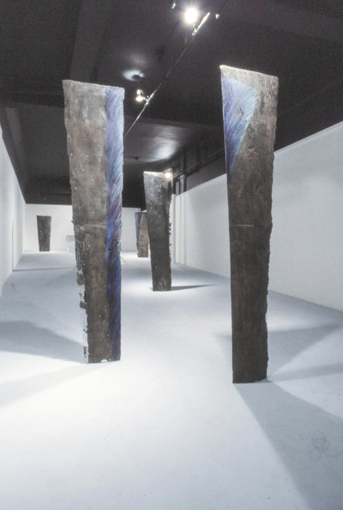 Several flat planks of cement fondu on wire stand vertically in a gallery space. They are glossy and well-lit, and have streaks of blue and purple along their flat edge.