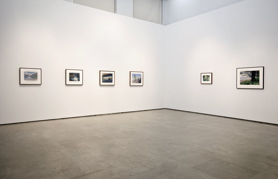 Six framed paintings of various sizes hang on either side of a corner gallery wall. On the left, there are four paintings of the same size. On the right wall, two paintings of different sizes hang.