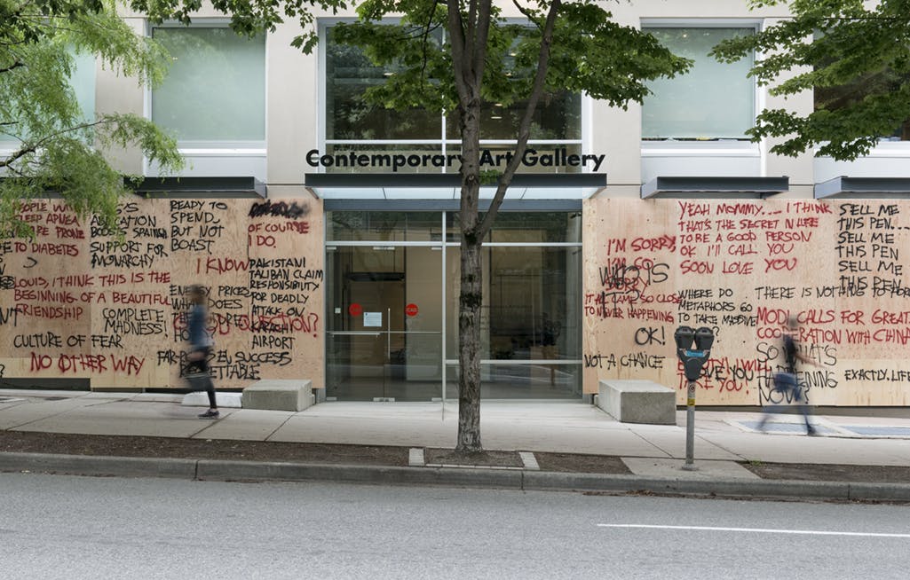 Multiple plywood boards installed on CAG’s facade blocking all the windows. Overlapping spray-painted words in red and black cover the boards. 