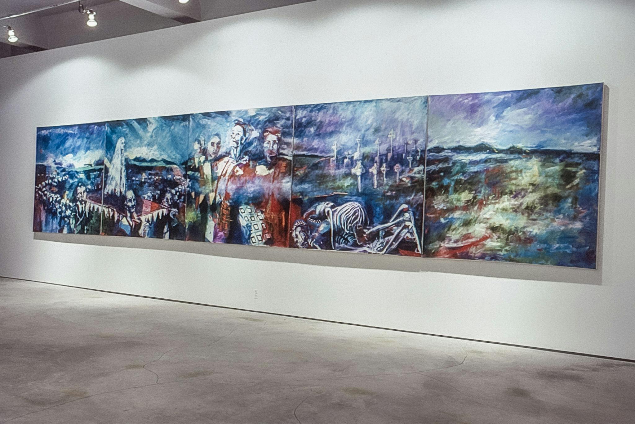 A large painting with greens, purples, reds, and blues, takes up most of a wall in a gallery space. The painting is made of 5 separate canvases, and depicts several skeletons and ghostly people.