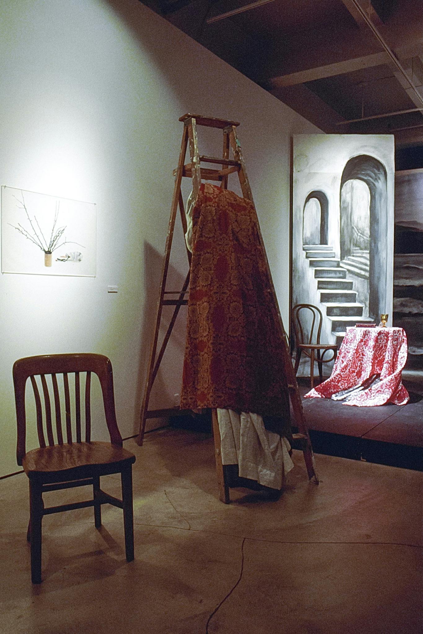 A tall wooden step ladder is placed in the middle of a gallery space. A yellow and red curtain, with a white layer underneath, are folded and hung from one of the ladder steps. 