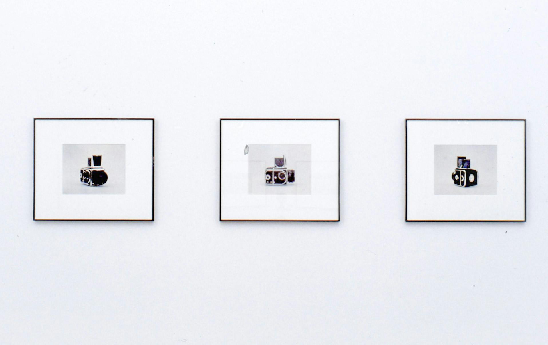 An installation image of three coloured photographs on a wall. These photographs depict different sides of an old black camera.