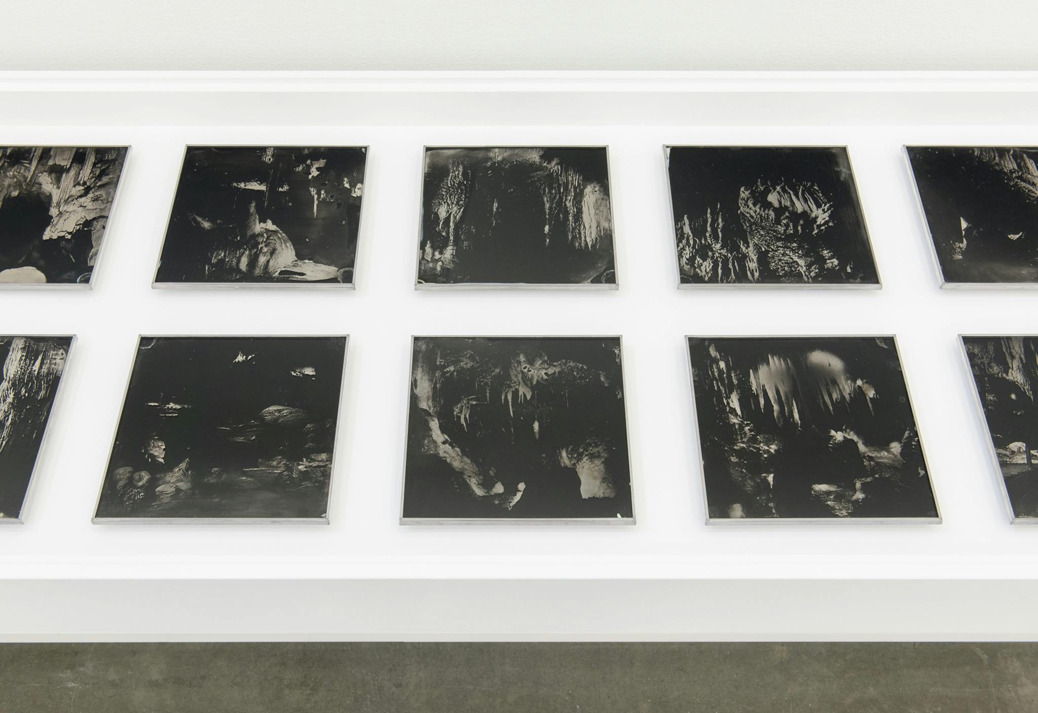 Ten photographs are visible on a display vitrine in a gallery. The square, low-exposure images show the inside of caves. The images reveal various stalactites and stalagmites formations.