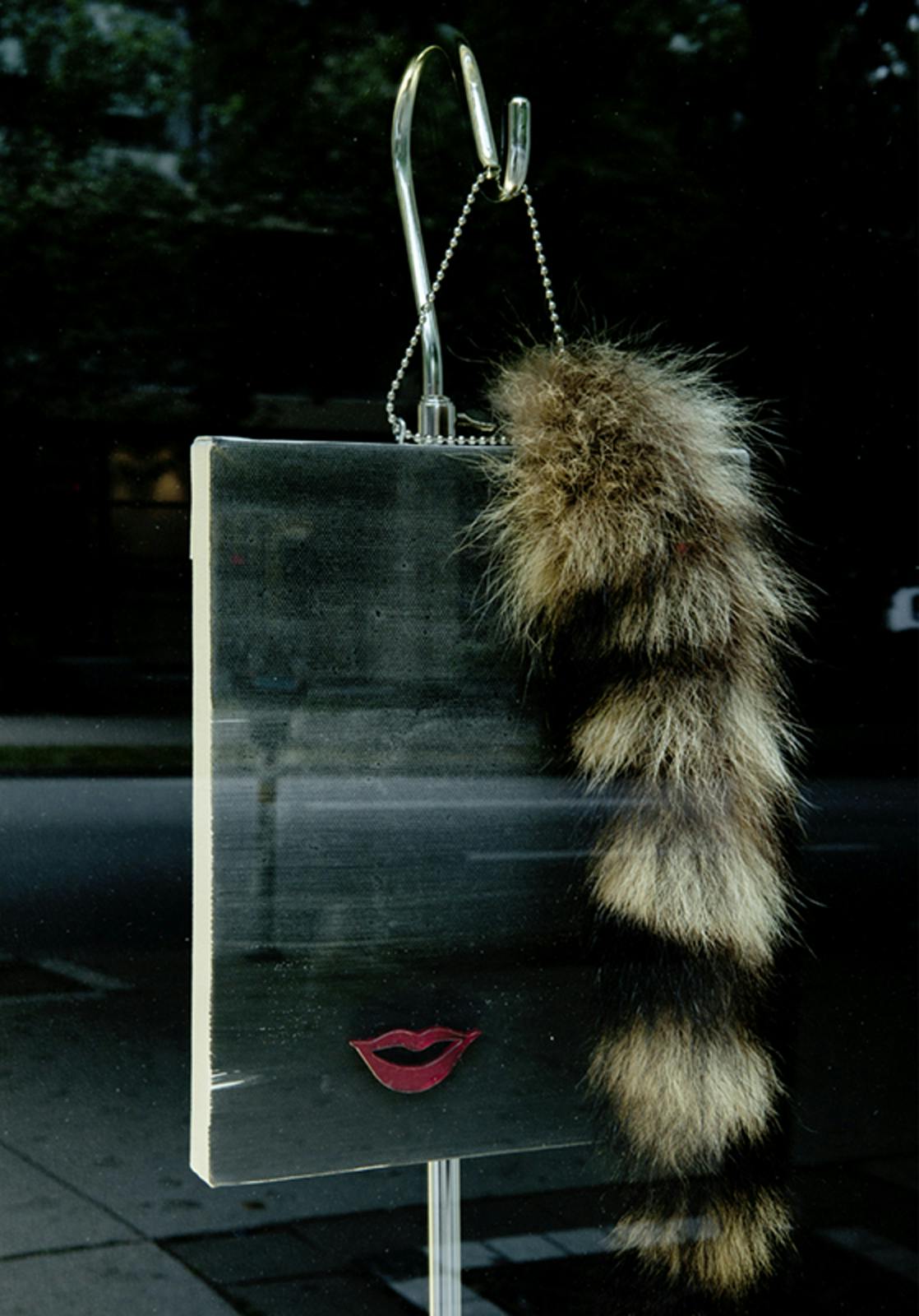 Josephine Meckseper’s sculpture installed in one of CAG’s facade windows. A fox tail ornament hangs from a silver, free-standing coat rack. A mirror is attached to the rack.