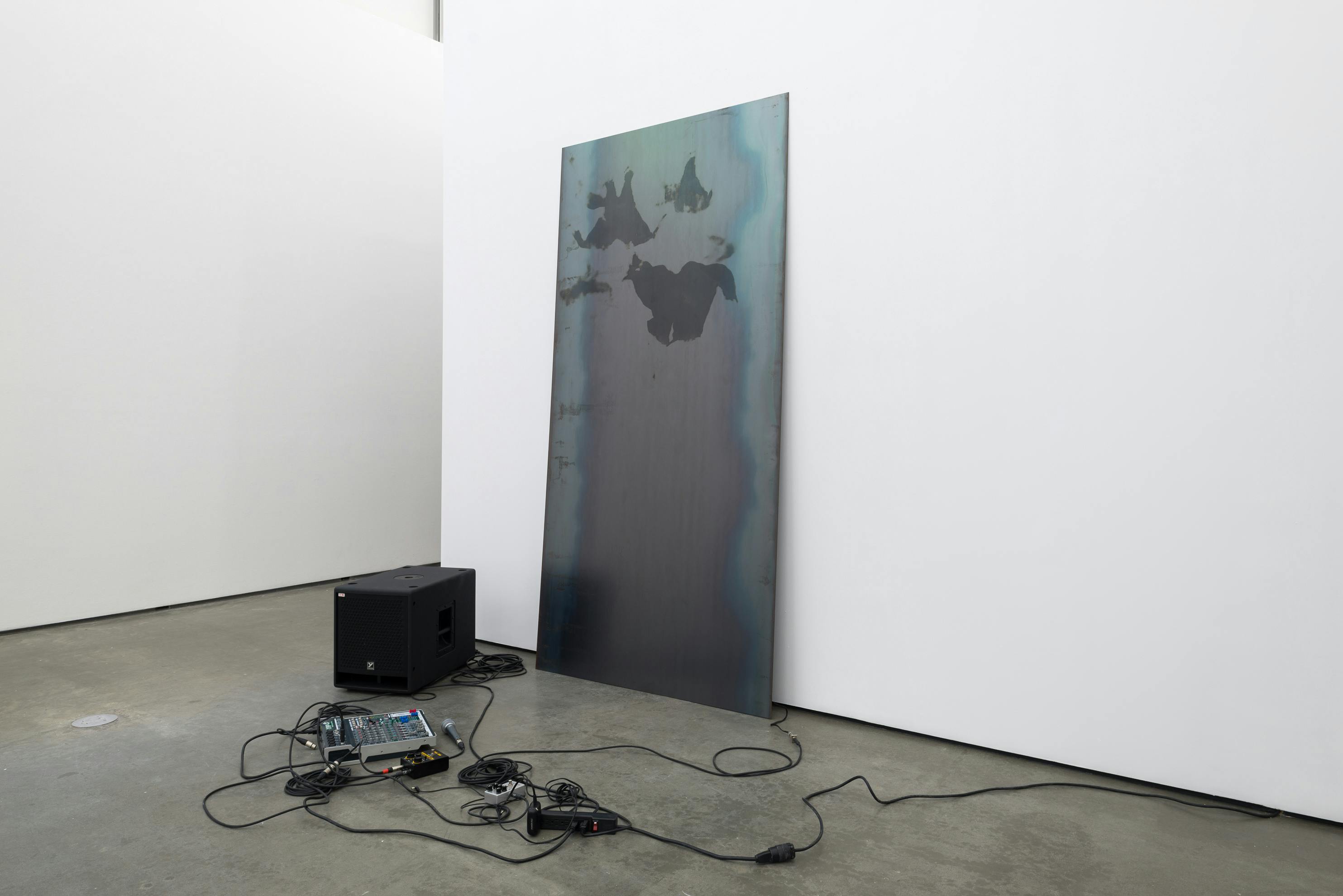 One large rectangular steel sheet leans against a wall of a gallery space. The sheet is marked with grease prints. On the floor surrounding the sheet, a microphone, speaker, mixer and patch cords sit.
