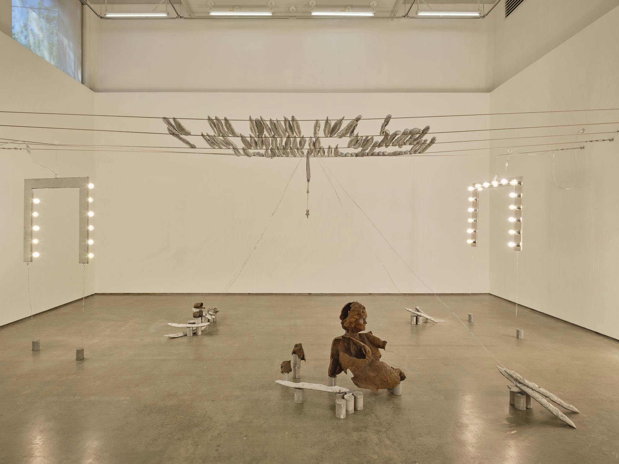 A wide view of several sculptures hanging from wire: magic wands, vanity lights and silver baguettes. A bronze sulpture of a partial human figure sits among aluminum cans and painted baguettes scattered across the floor.