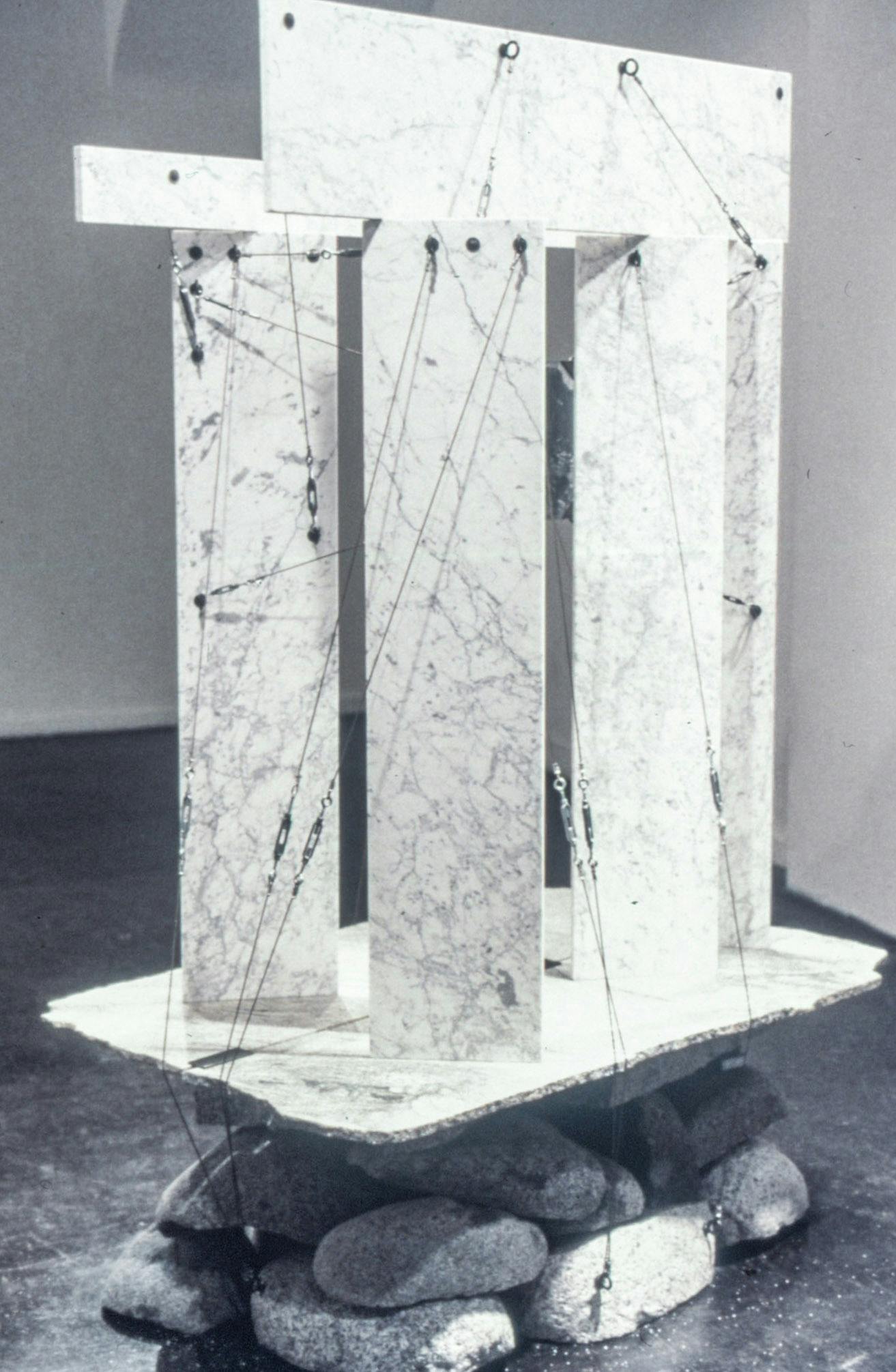 A closeup of a sculpture in a gallery. It is made of several marble slabs stacked vertically and horizontally, resting on a pile of large rocks. The pieces are attached by metal wire and hardware. 