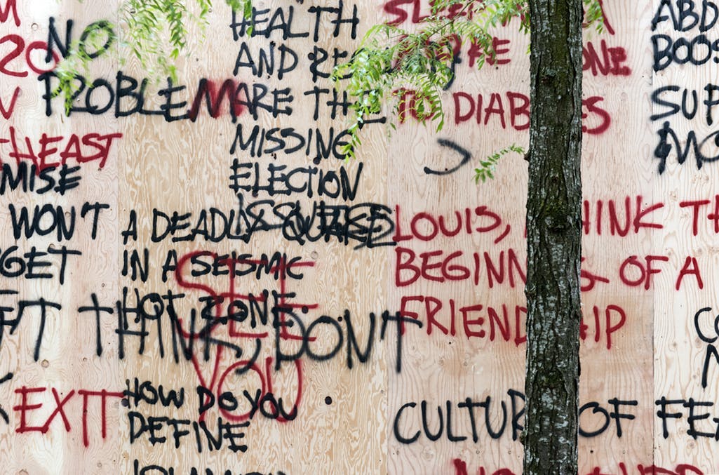 Detail image of plywood boards with multiple overlapping words in red and black spray paint. One phrase reads, “A DEADLY QUAKE IN A SEISMIC HOT ZONE.”