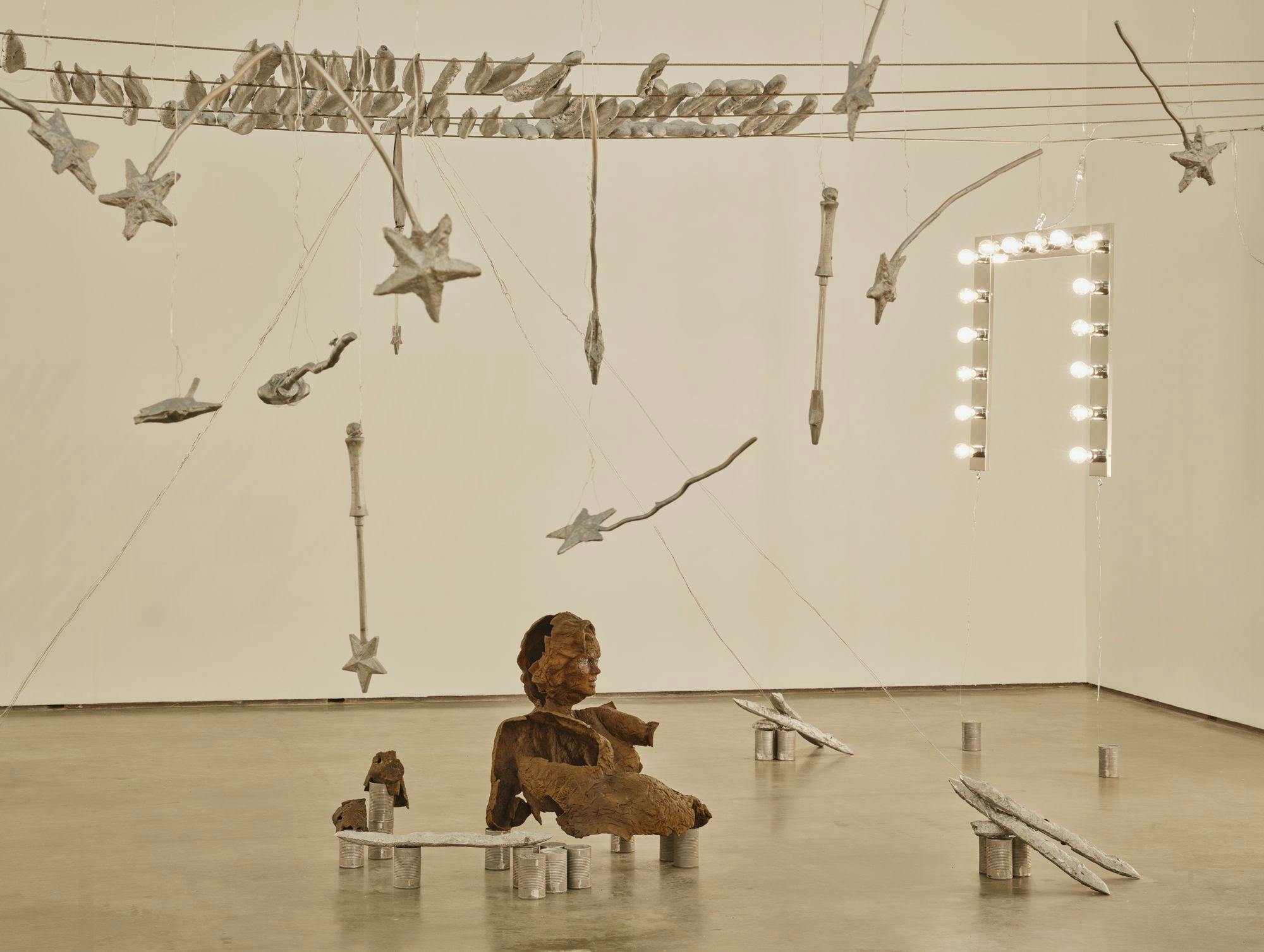 A wide view of several sculptures hanging from wire: magic wands, vanity lights and silver baguettes. A bronze sulpture of a partial human figure sits among aluminum cans and painted baguettes scattered across the floor.