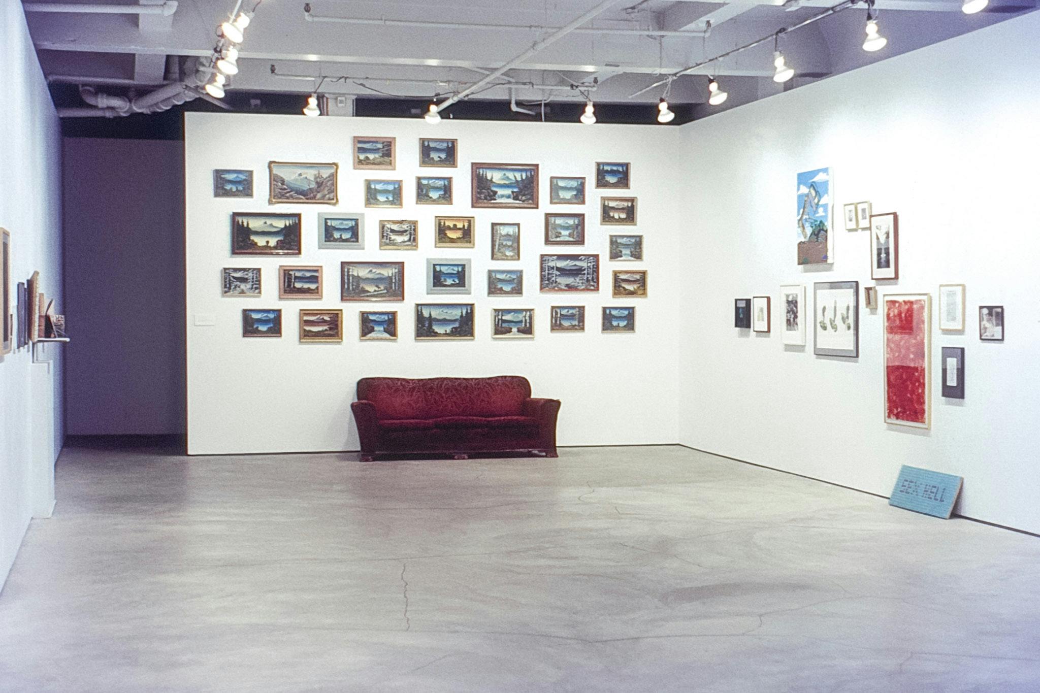 Three white walls and a shadowy hallway in a gallery. Dozens of artworks are mounted on the walls. The works on the back wall are landscapes, with a faded red velvet couch in front of them.
