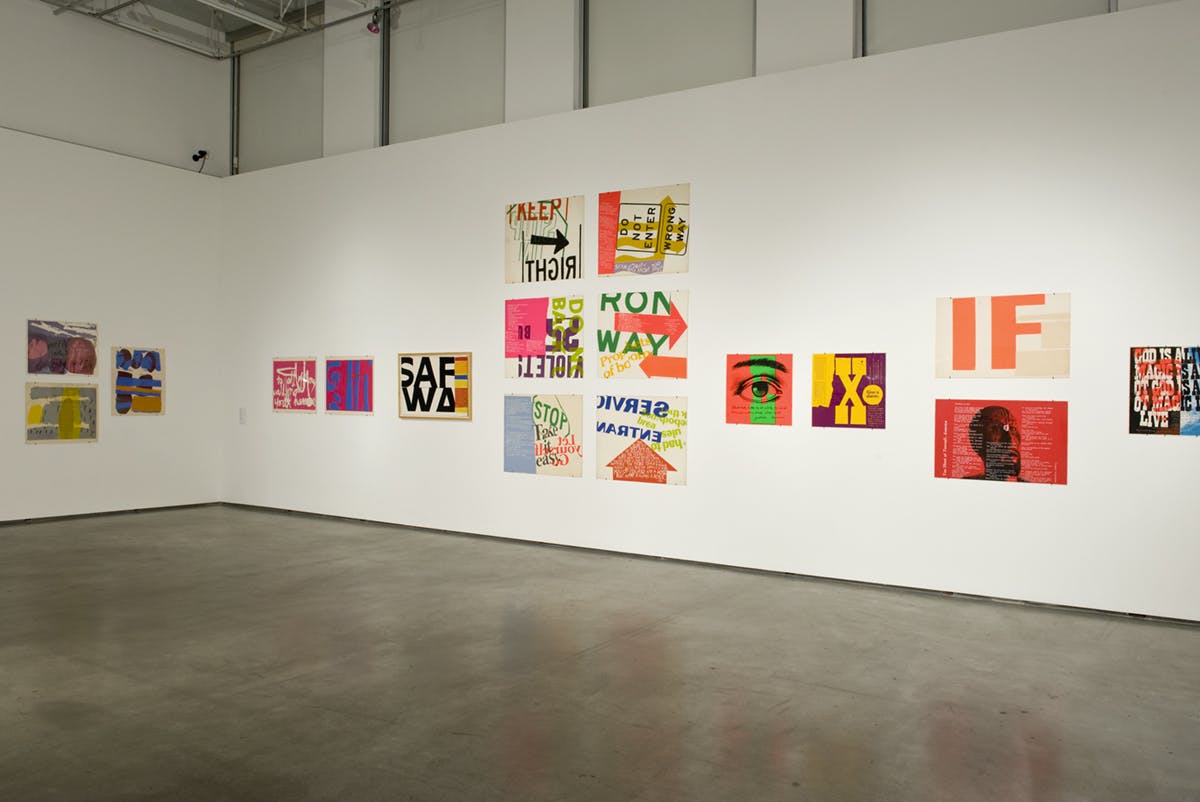 Installation view of Corita Kent’s colourful sign artworks mounted on white gallery walls. A poster of a large realistically drawn eye is installed next to the yellow and purple sign.